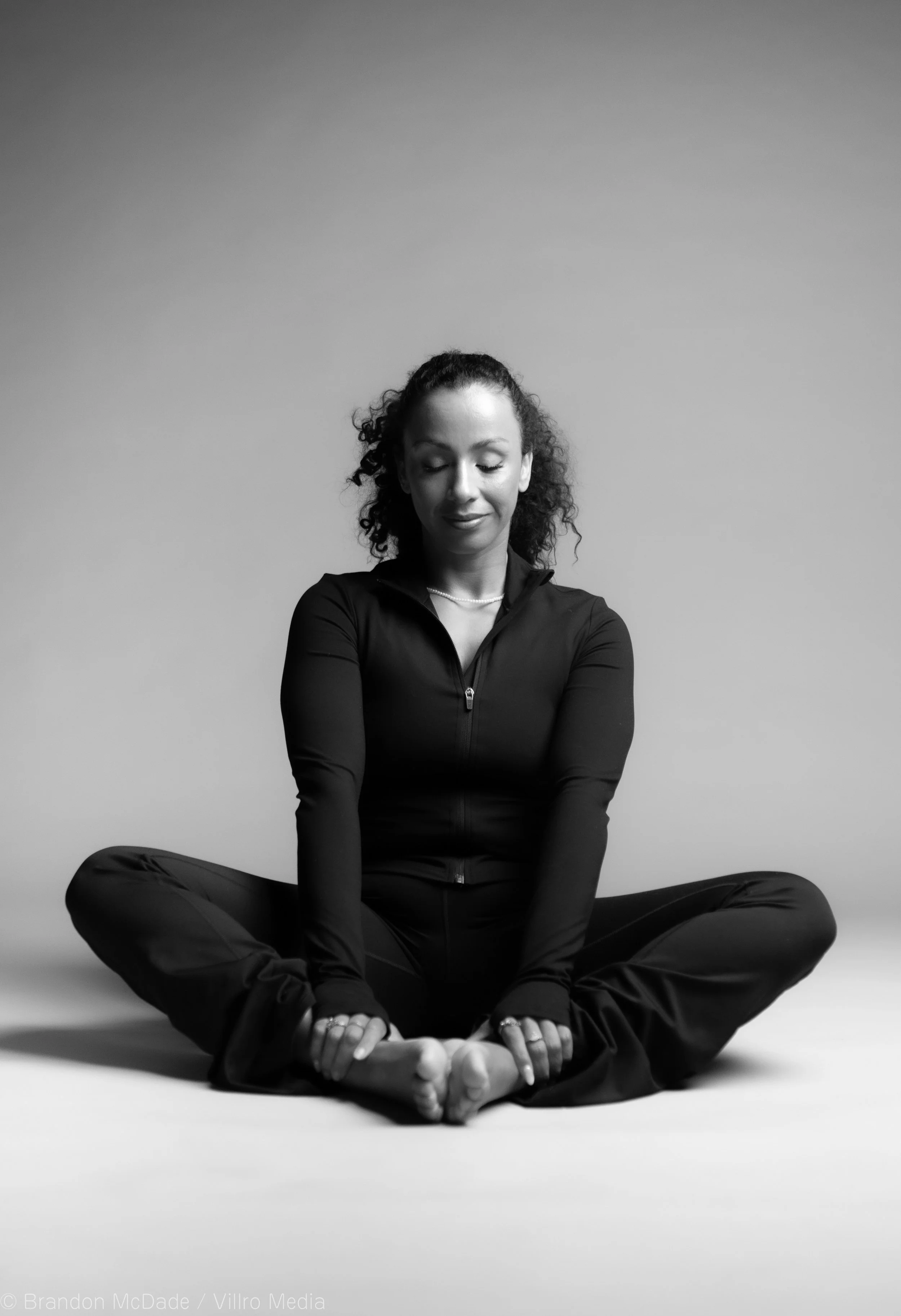 A woman is sitting cross-legged, meditating with eyes closed in a black athletic outfit in a studio setting.