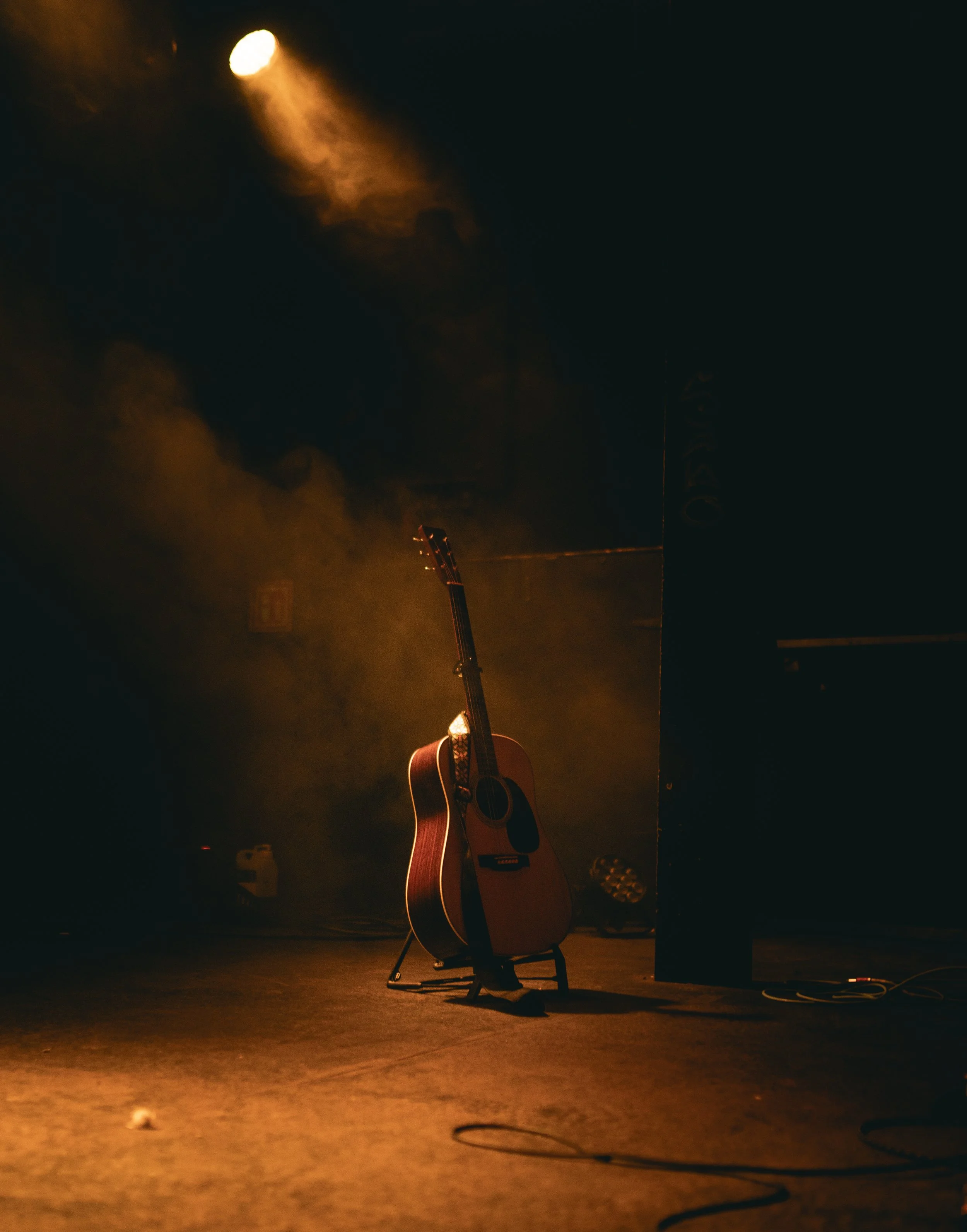 An acoustic guitar on a stand on a dimly lit stage, with a spotlight shining from above and smoke in the background.