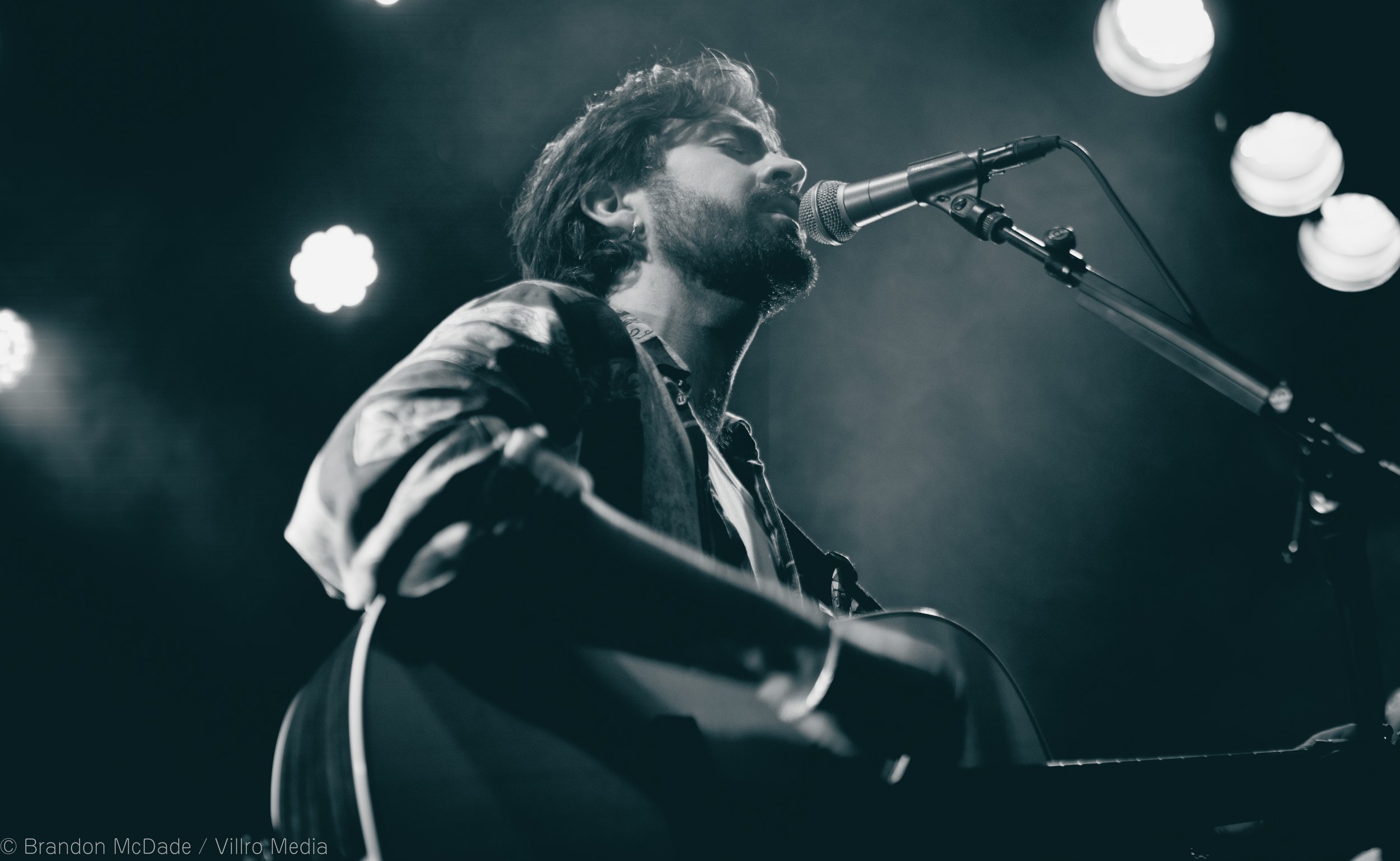 A man playing an acoustic guitar and singing into a microphone on stage, with stage lights overhead, in black and white.
