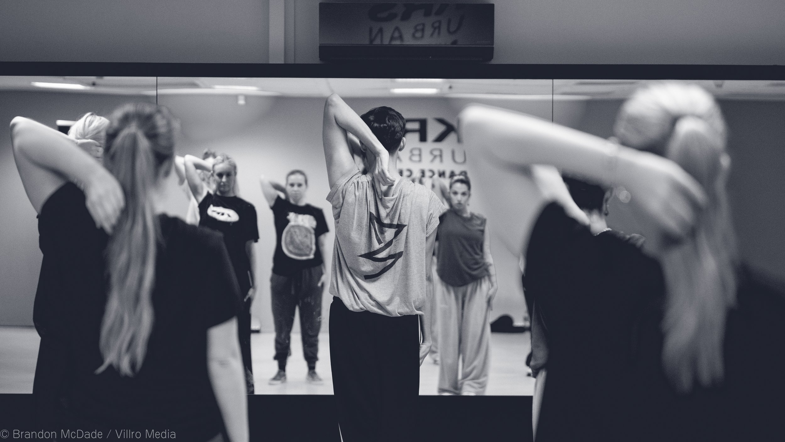 A dance or fitness class in a studio, with students stretching or warming up in front of a mirror, and an instructor visible in the reflection. The photo is in black and white.