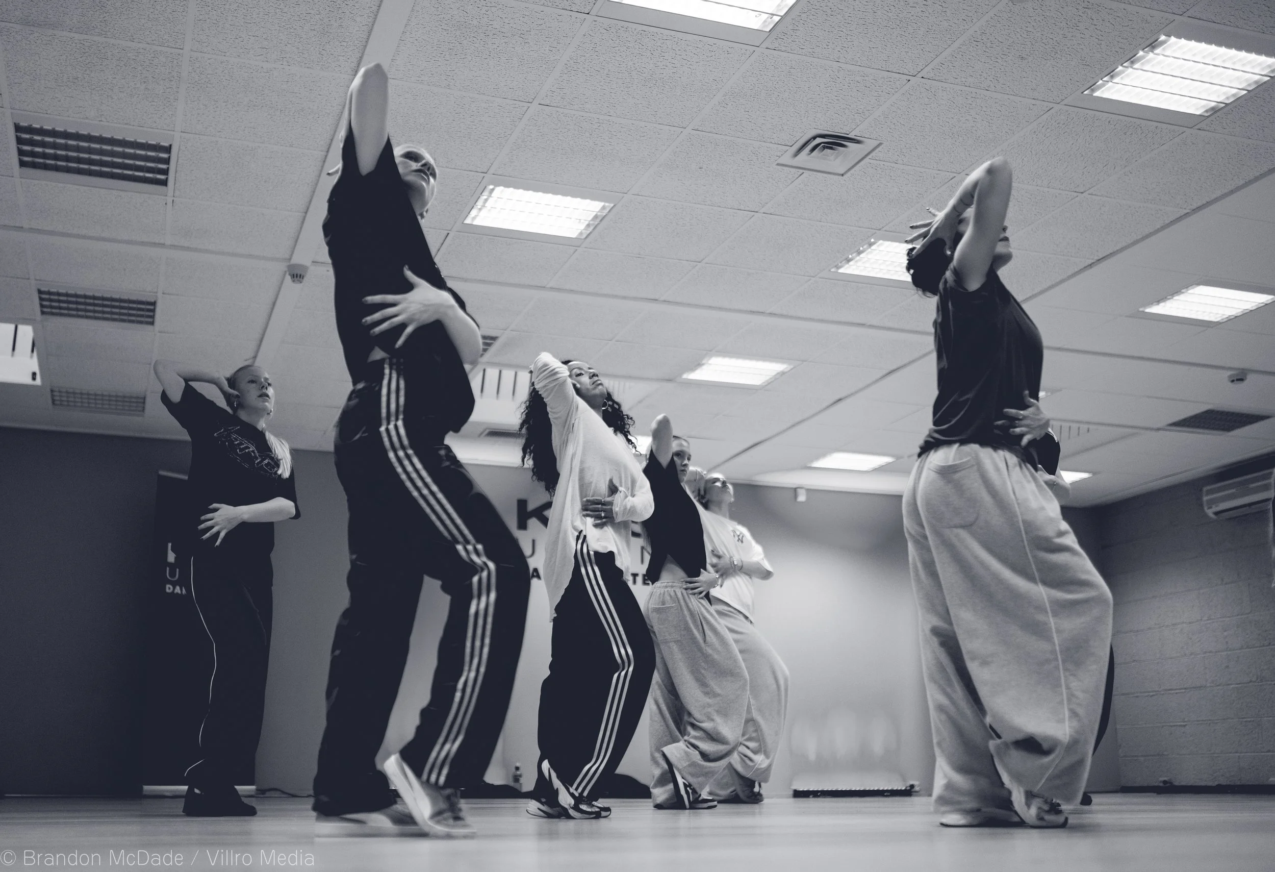 Group of dancers rehearsing in a dance studio, practicing a coordinated routine. The dancers are dressed in casual comfortable clothing and are in various poses, with some having their hands on their hips or heads.