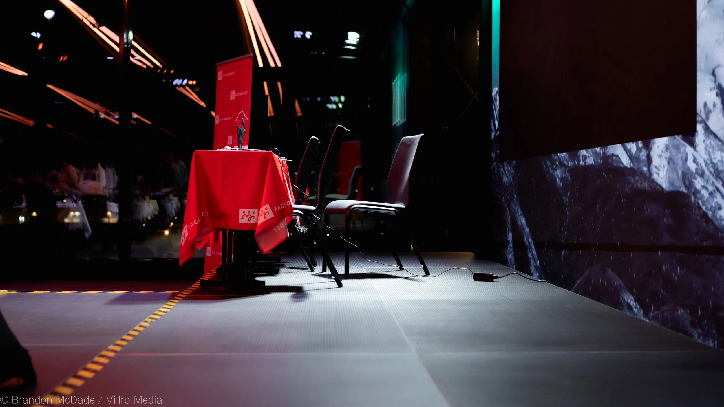 A stage with a round table draped with a red cloth, three chairs, a microphone, and some cables in front of a dark backdrop with projected images.