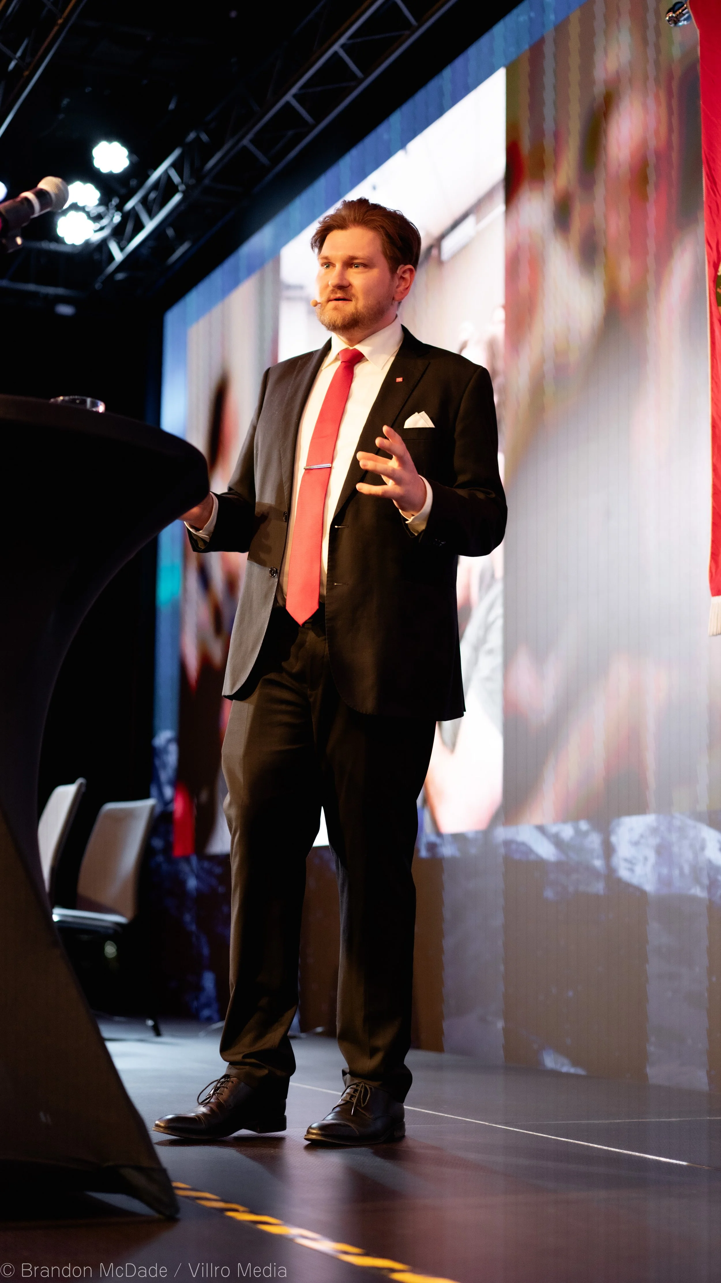 Man in formal suit and red tie giving a presentation on stage with large screen behind him.