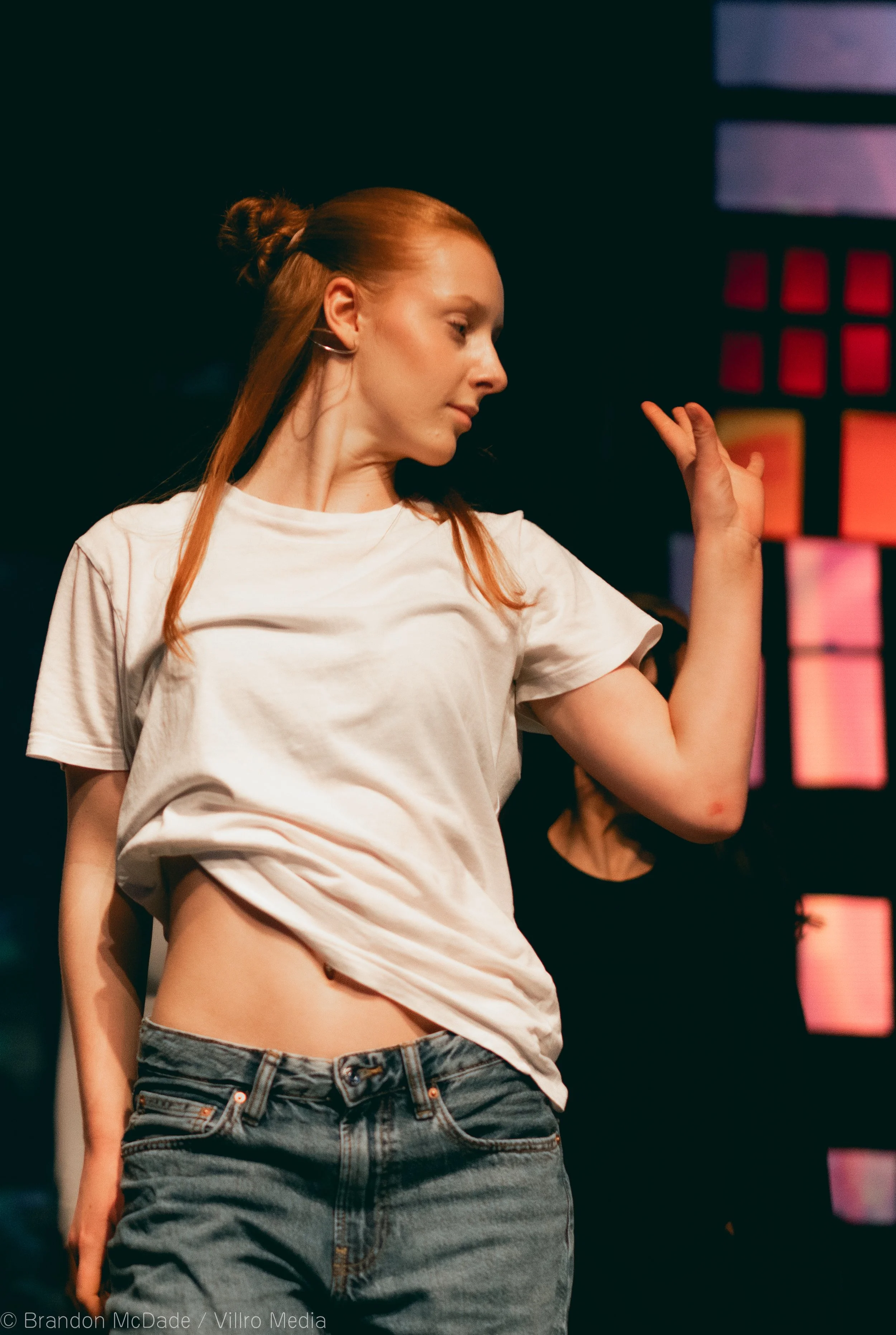 Young woman with red hair in a bun, wearing a white T-shirt and blue jeans, dancing against a colorful background.