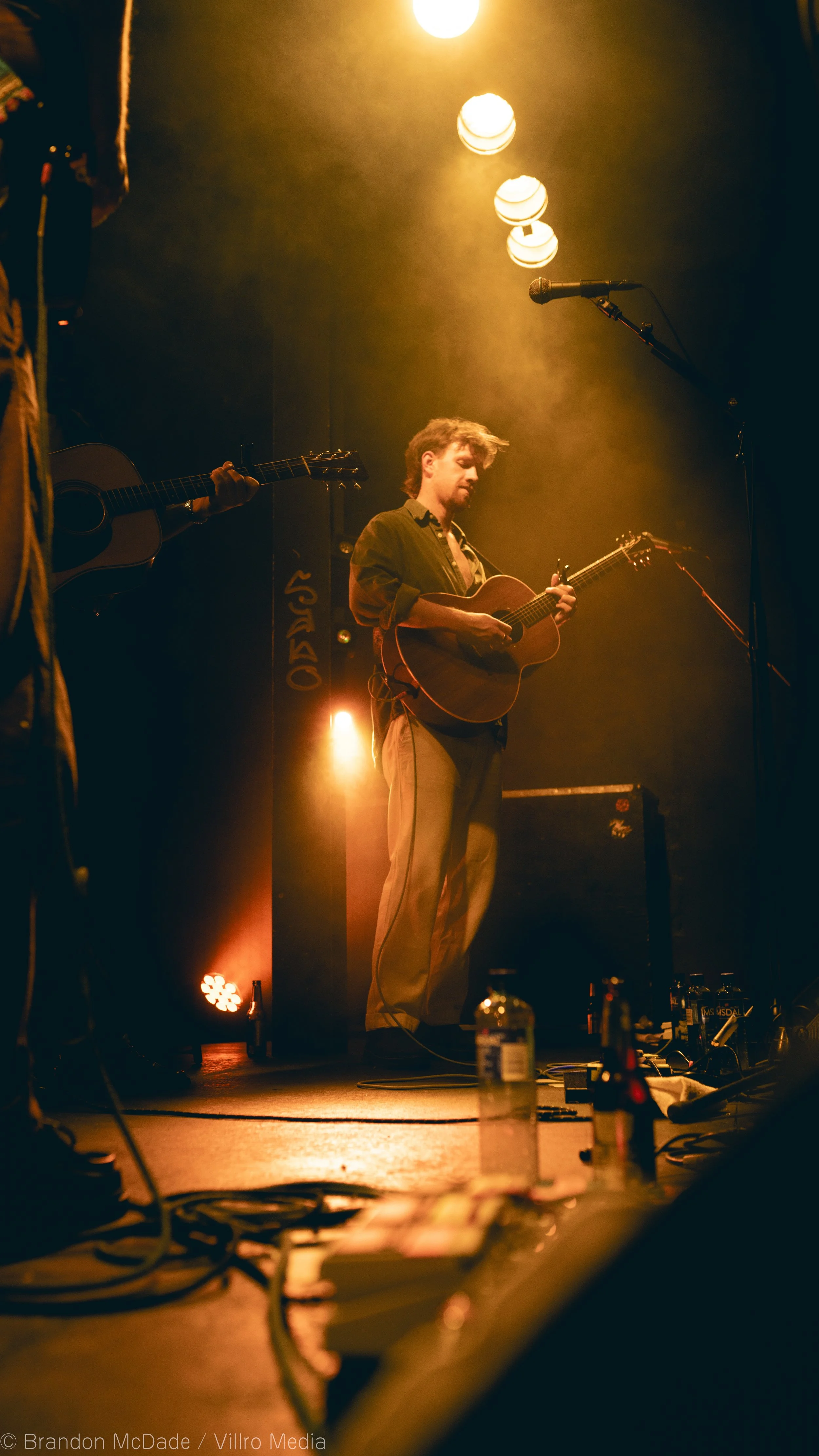 A musician on stage playing an acoustic guitar during a live performance, with stage lights illuminating the scene and some bottles and equipment on the floor.