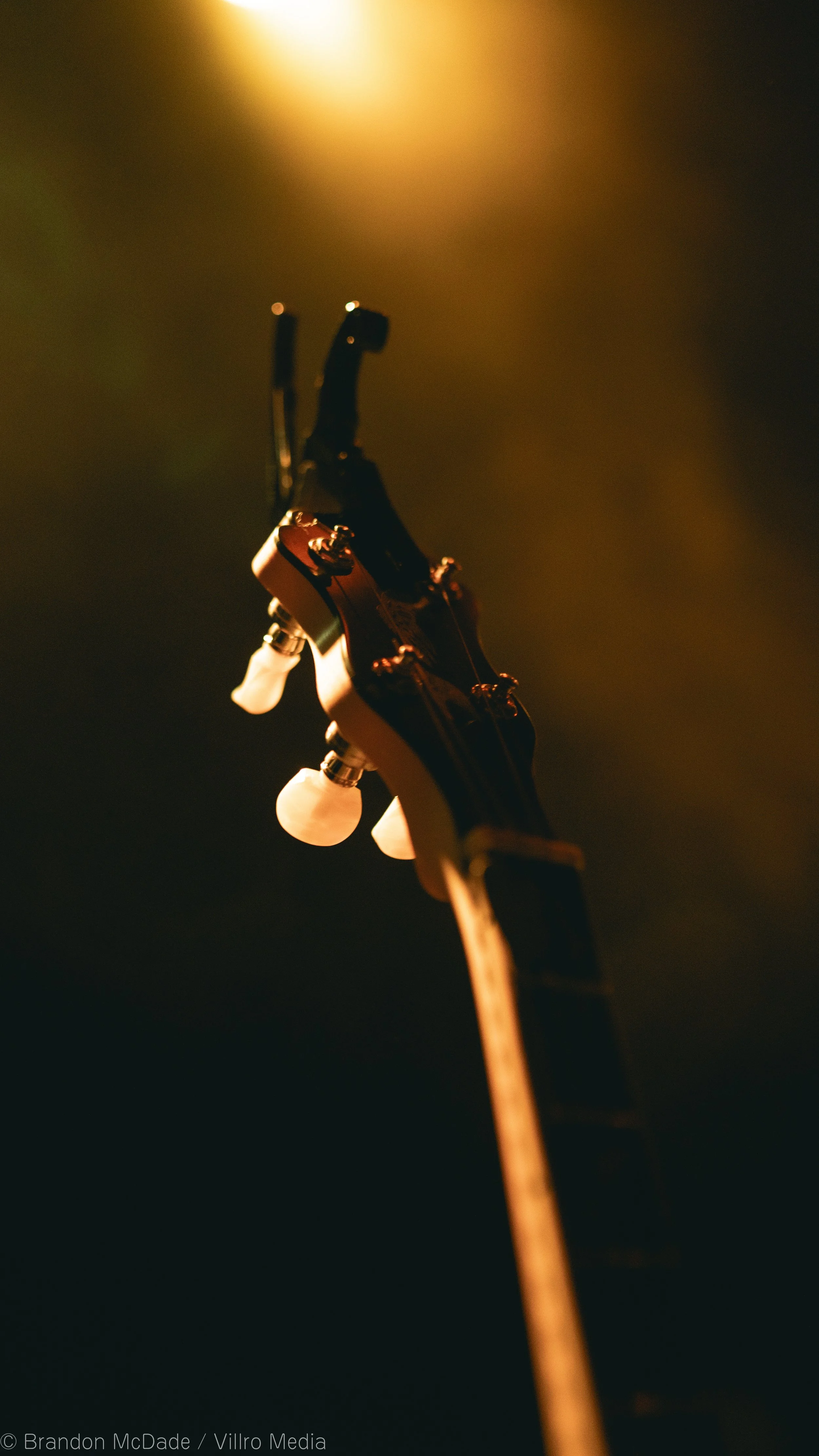 Close-up of a guitar headstock with tuning pegs, viewed from below in warm lighting.