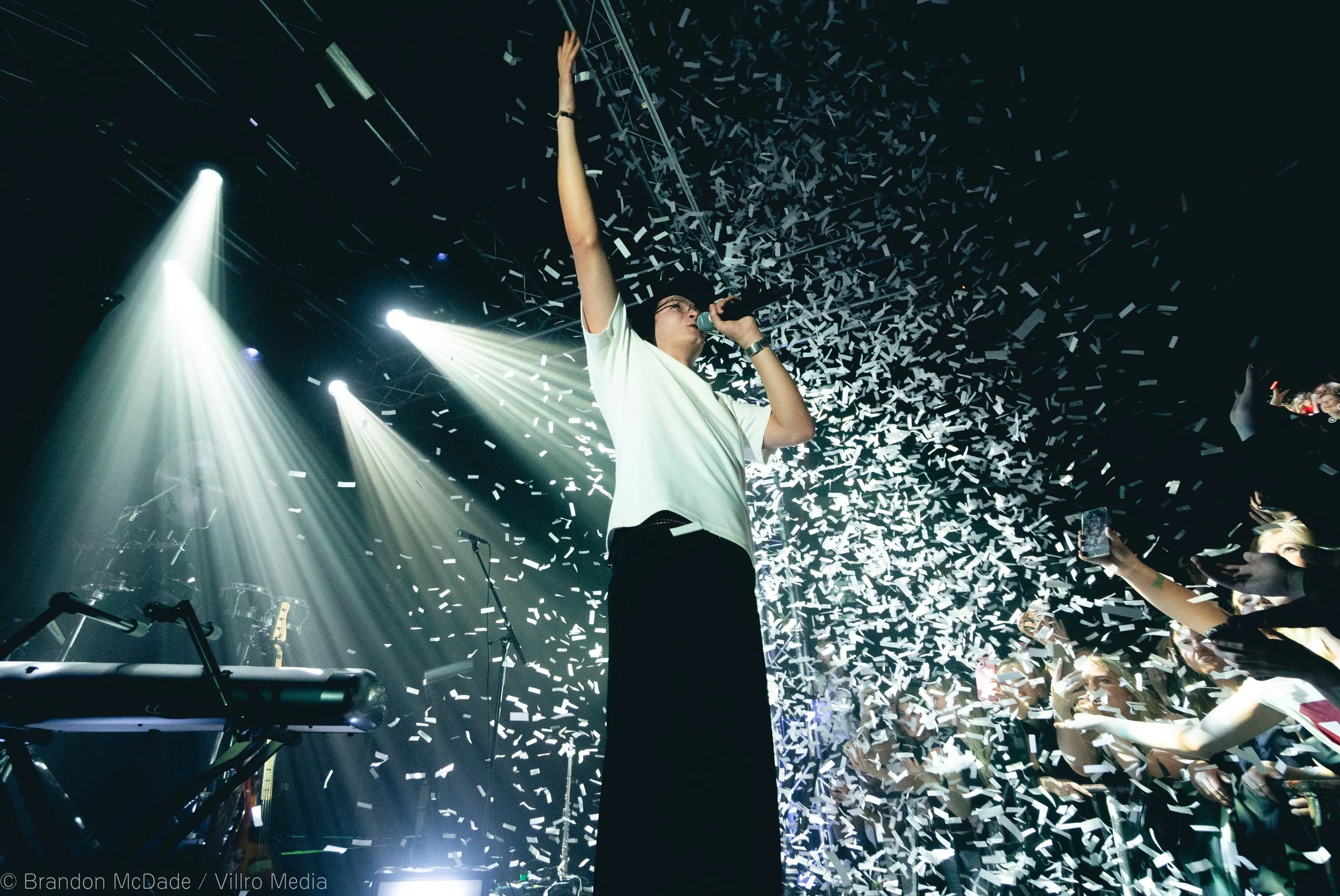 Performer on stage with white confetti falling, holding microphone, raising one arm, with bright stage lights and crowd taking photos.