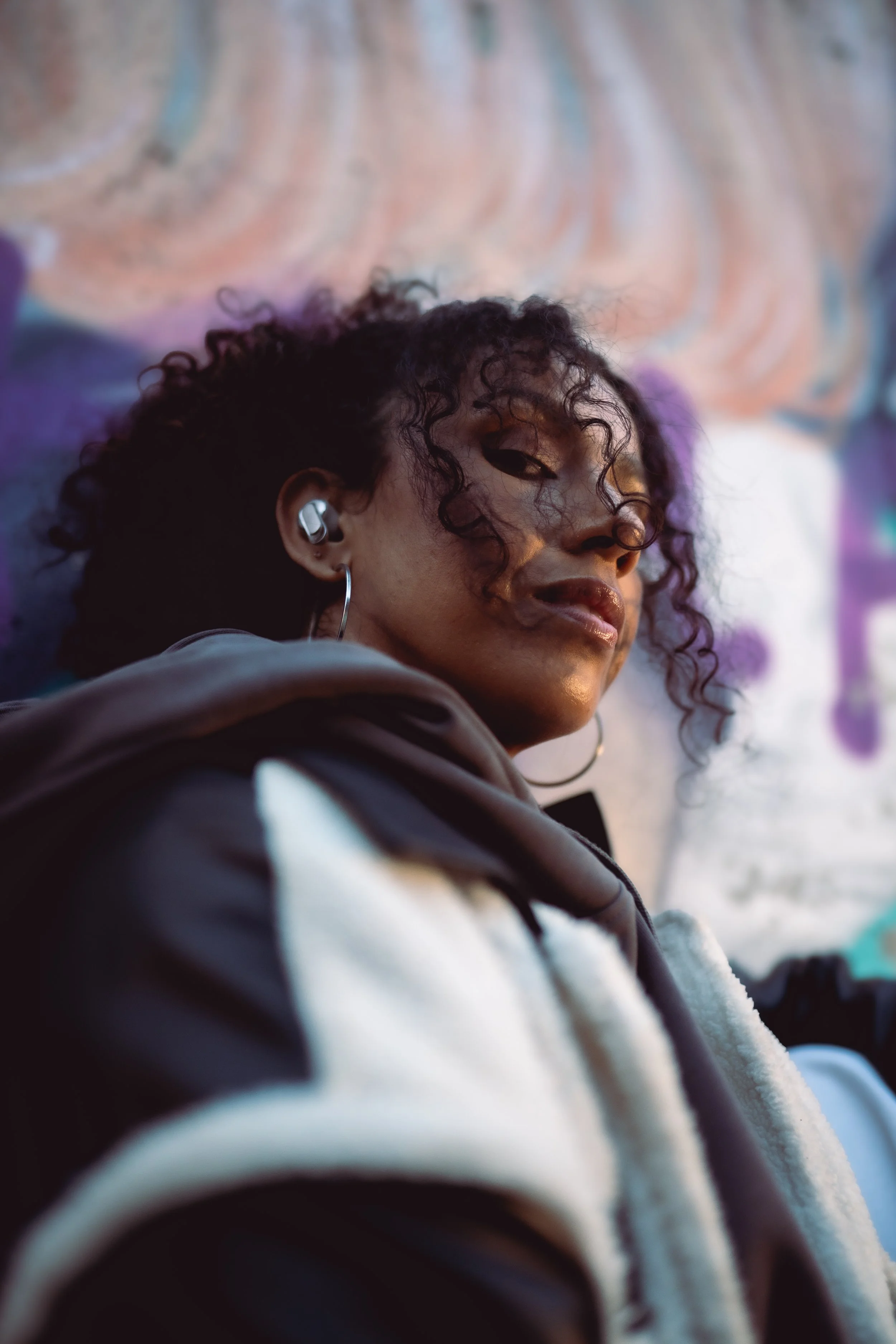A young woman with curly hair, wearing earbuds, resting against a colorful graffiti wall.