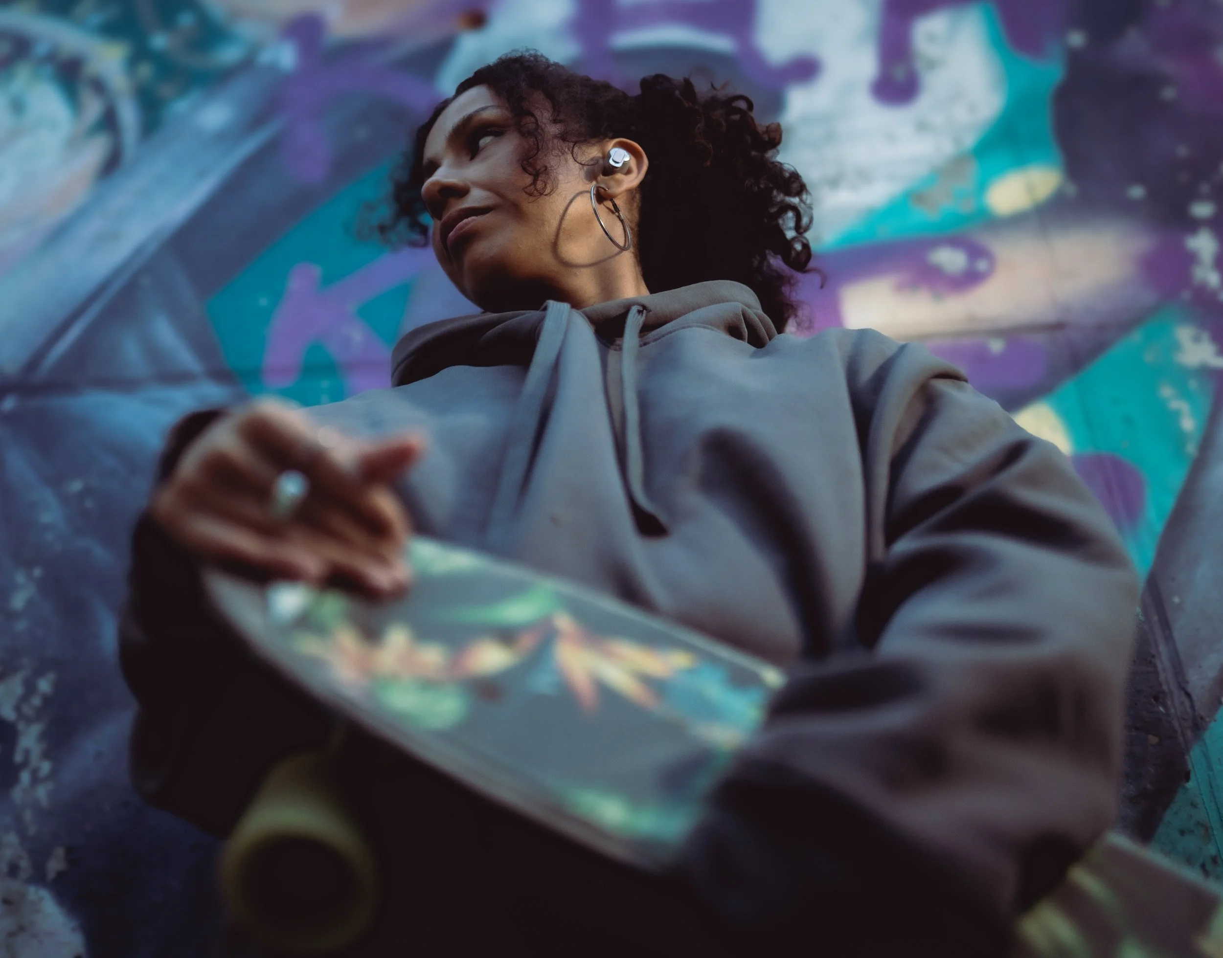 A woman with dark curly hair, wearing a hoodie, holding a skateboard, and standing in front of a graffiti-covered wall.