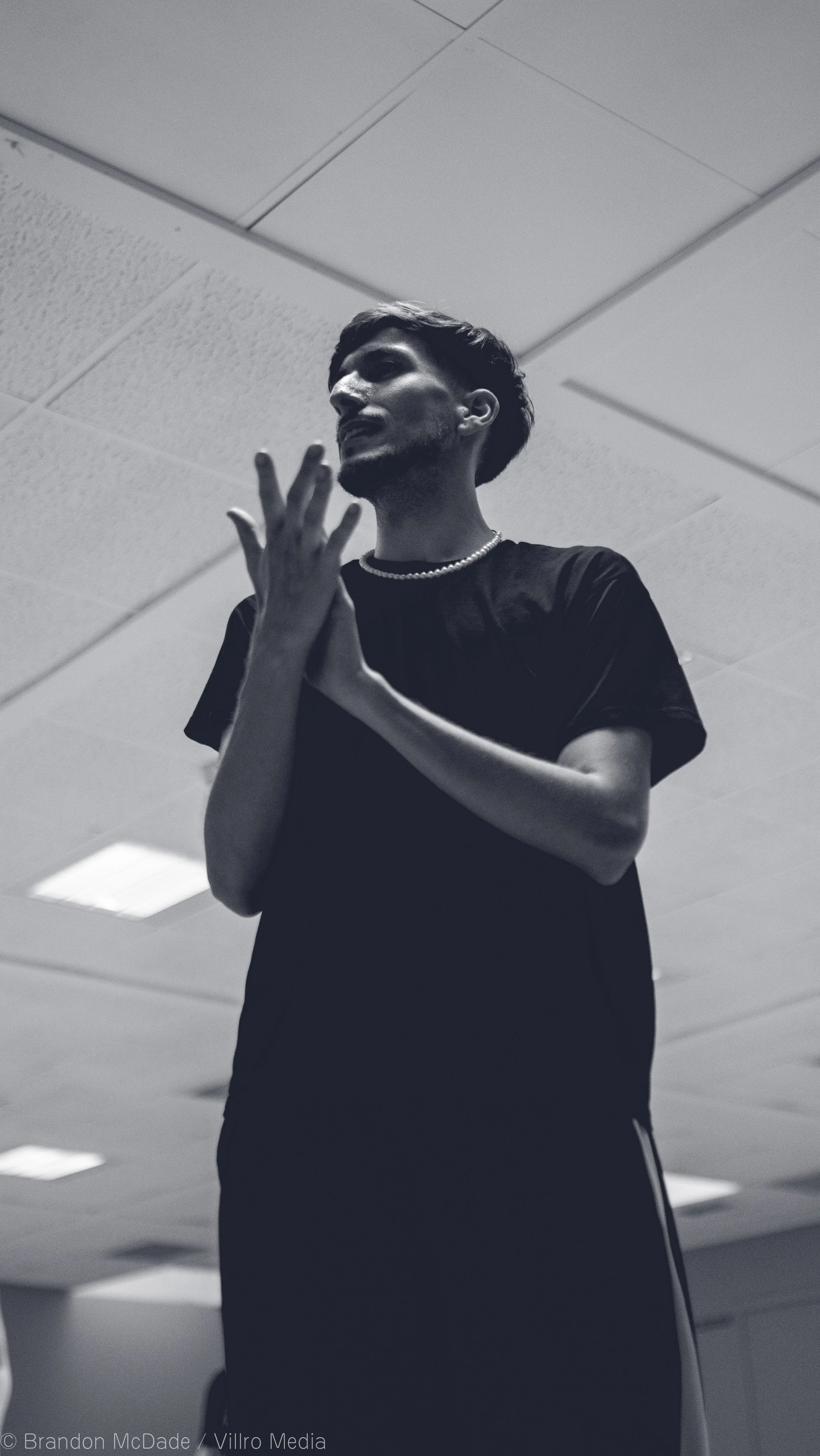 Black and white photo of a person with short hair, wearing a t-shirt, pearl necklace, and track pants, standing indoors with a ceiling with tiles above. They are holding their hands together in front of their chest and speaking or singing.