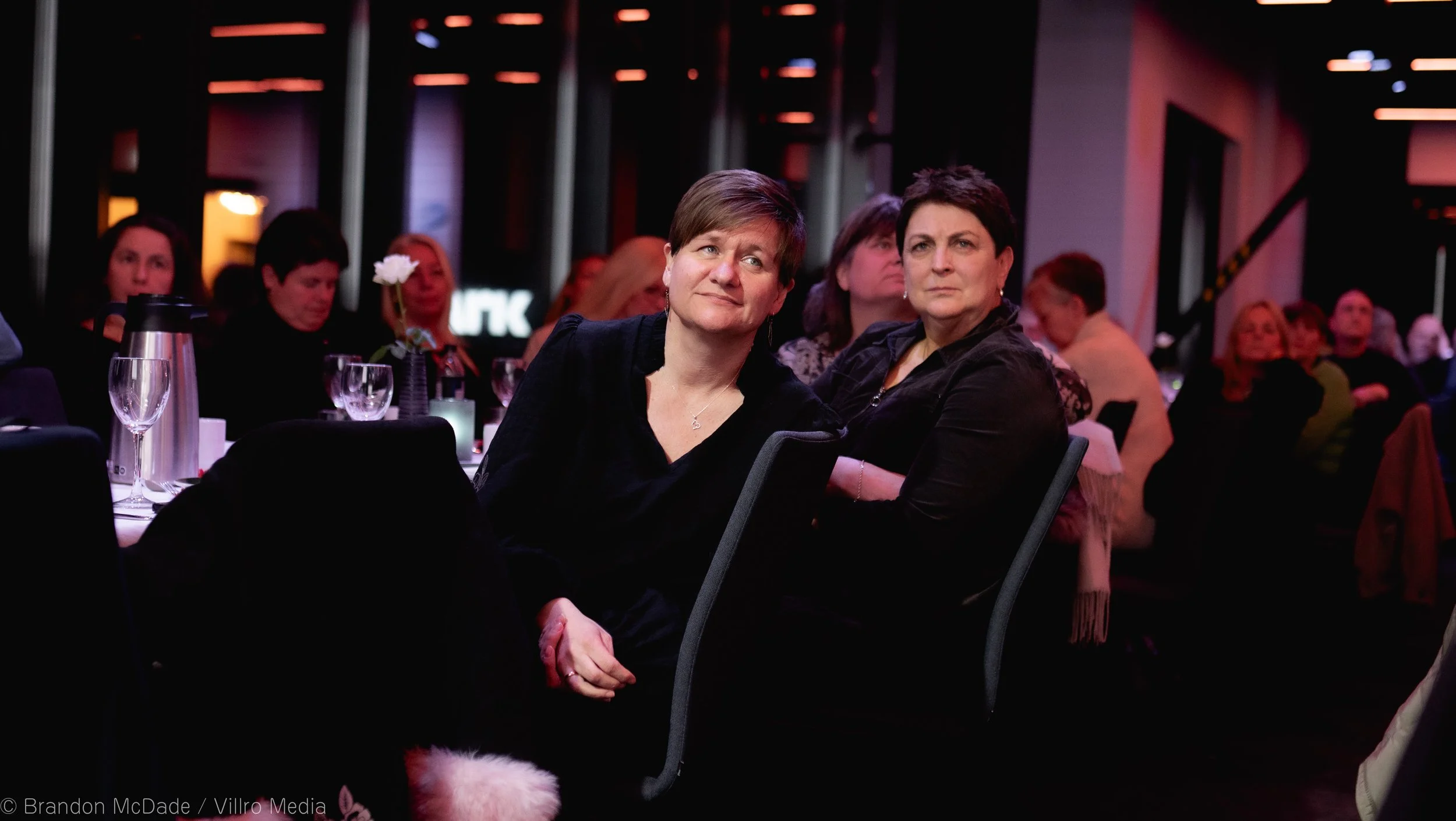 Two women sitting at a table during an indoor event, surrounded by other attendees, some with glasses and flowers on the table, with a dark background and colorful lighting.