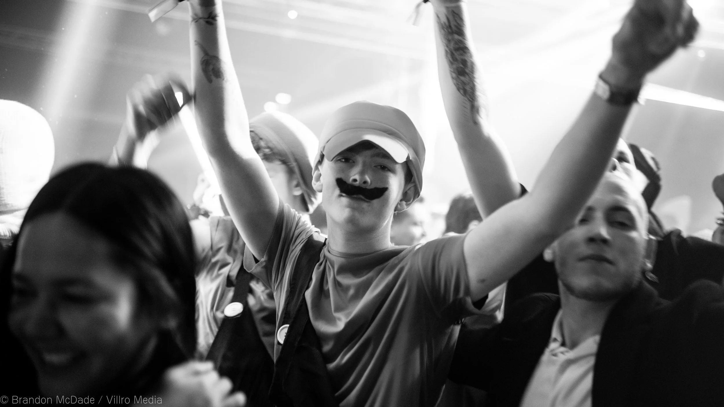 A group of people celebrating at a party, with a young person in the center wearing a hat and mustache makeup, raising their arms in excitement.