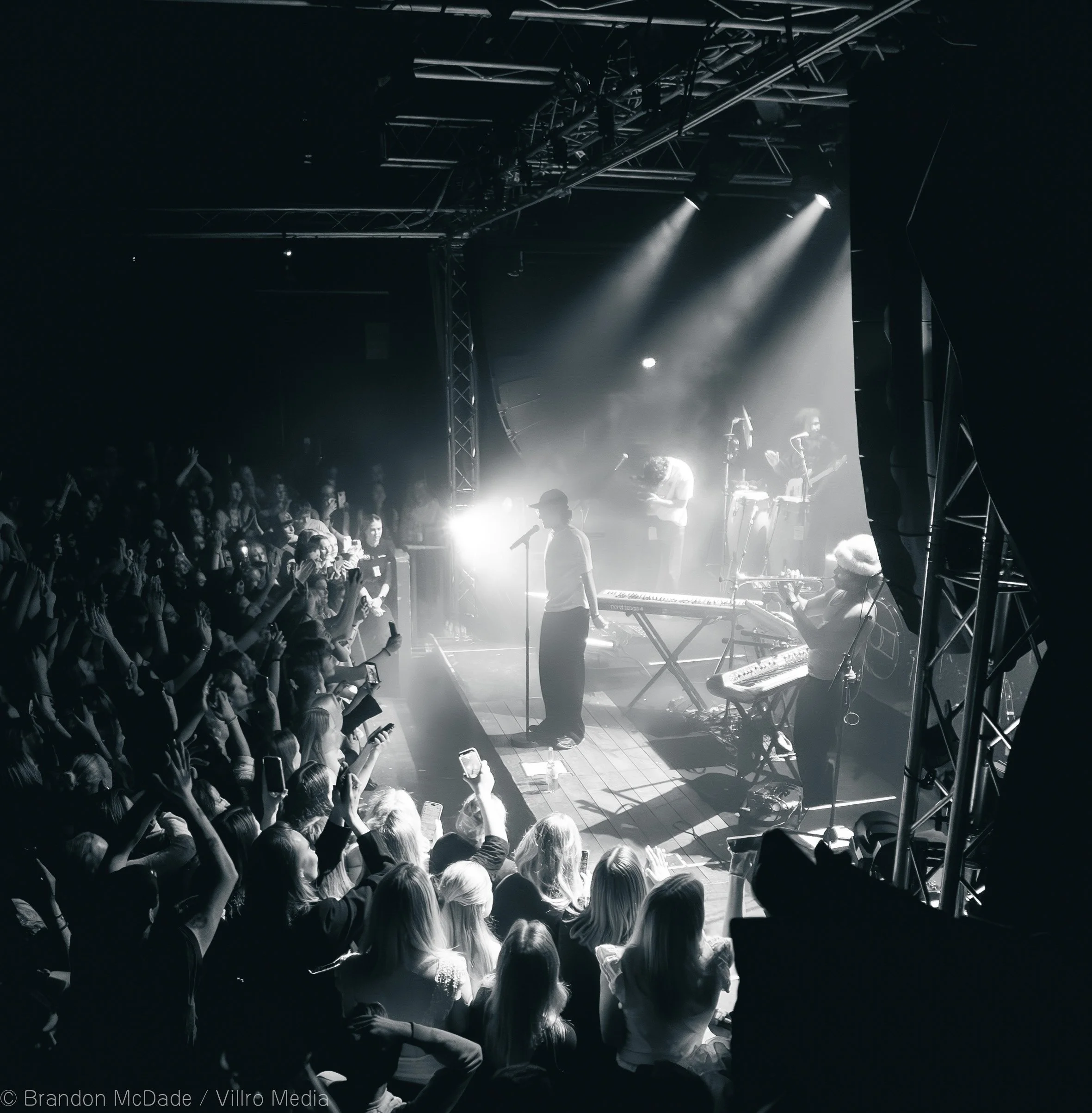Black and white photo of a live concert with a singer at the microphone in front of an audience, band members playing instruments on stage, and concert lighting.