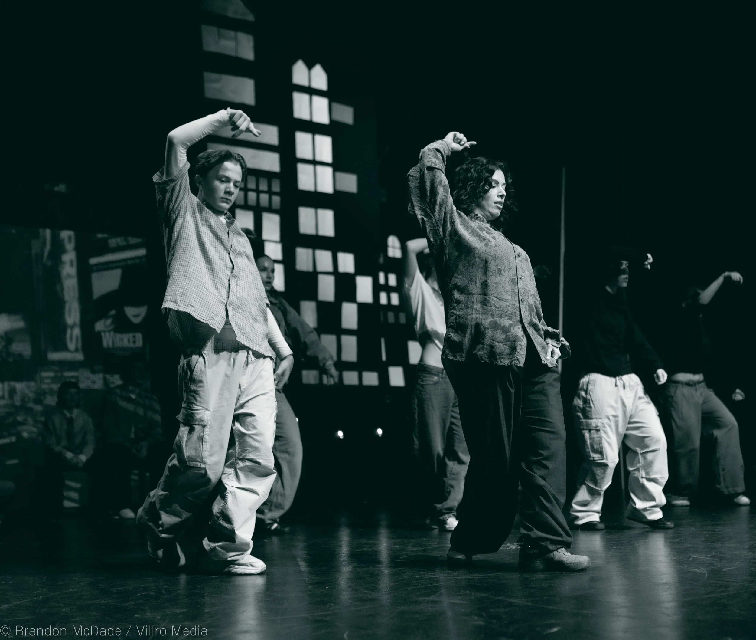 A group of dancers performing on a stage with a dark cityscape backdrop, captured mid-movement.