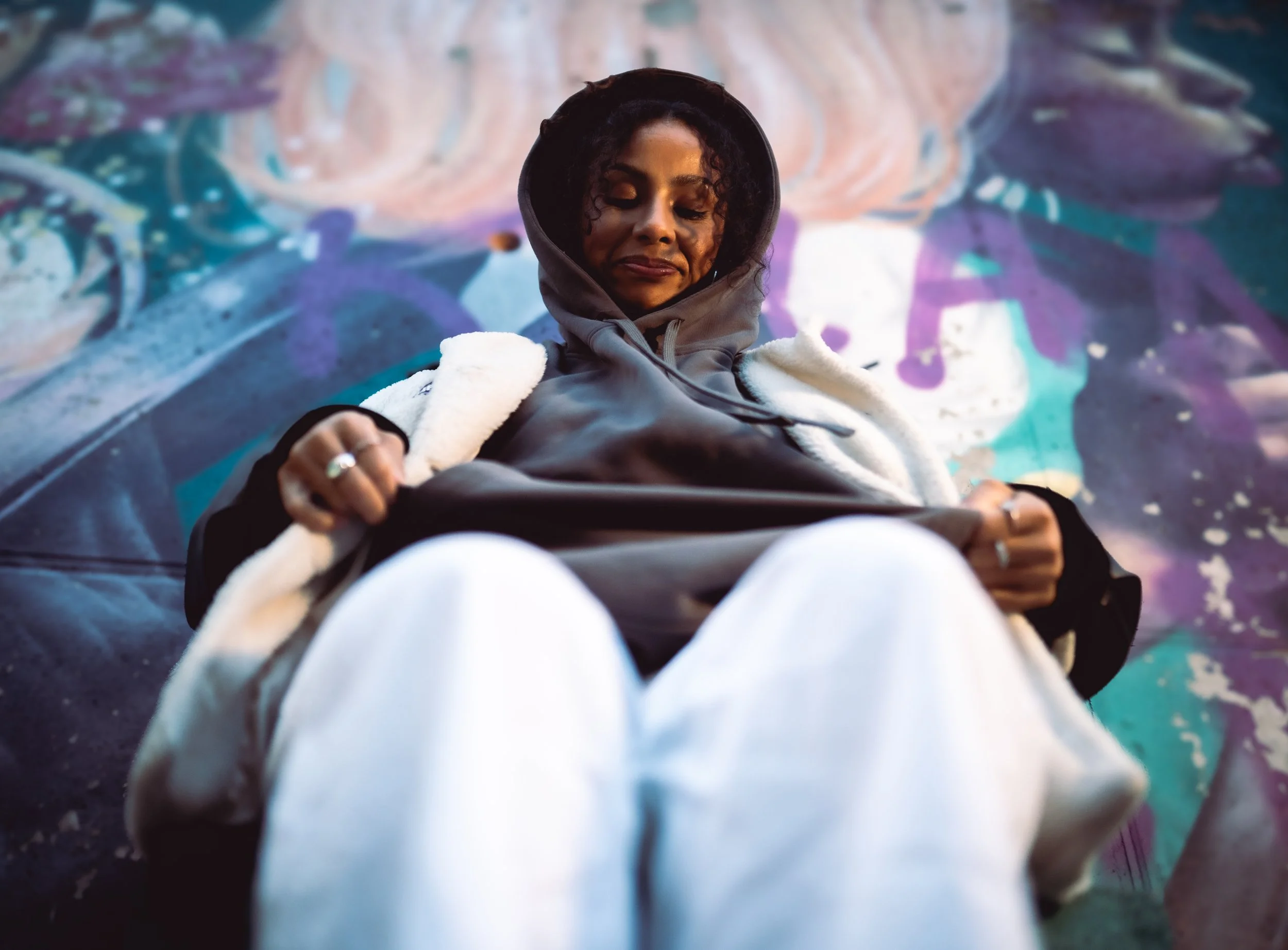 A woman in a gray hoodie and white pants sitting against a colorful graffiti wall, looking down and adjusting her clothing.