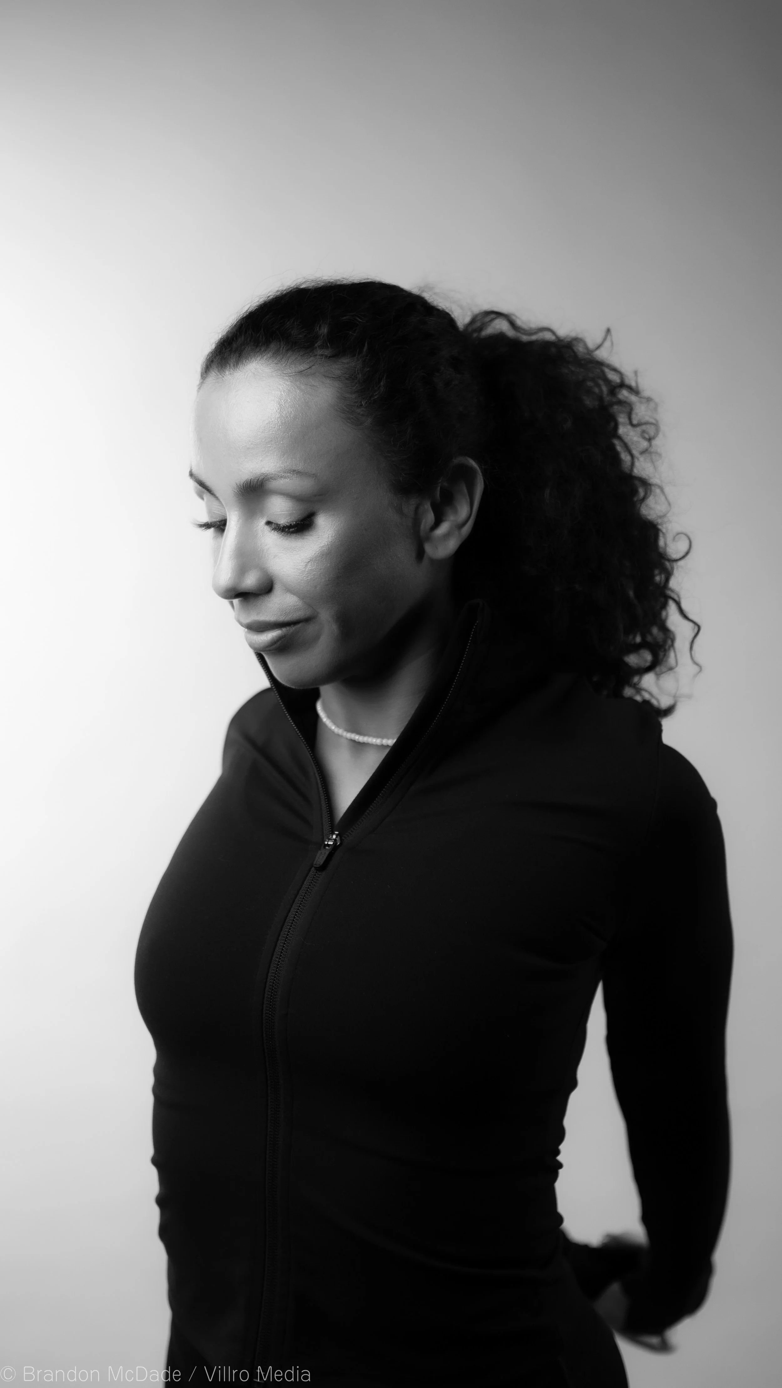 Black and white portrait of a woman with curly hair pulled back, wearing a zip-up jacket and a pearl necklace, with her head tilted down and eyes closed.