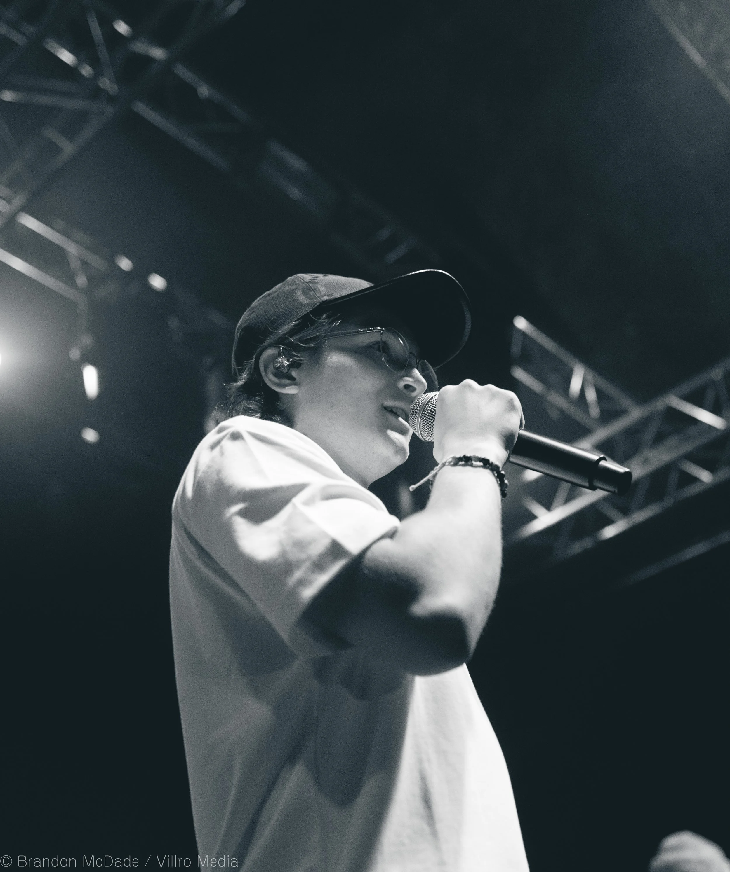 A young man standing on stage, holding a microphone, wearing glasses, a cap, and casual clothing, possibly performing or speaking at an event.