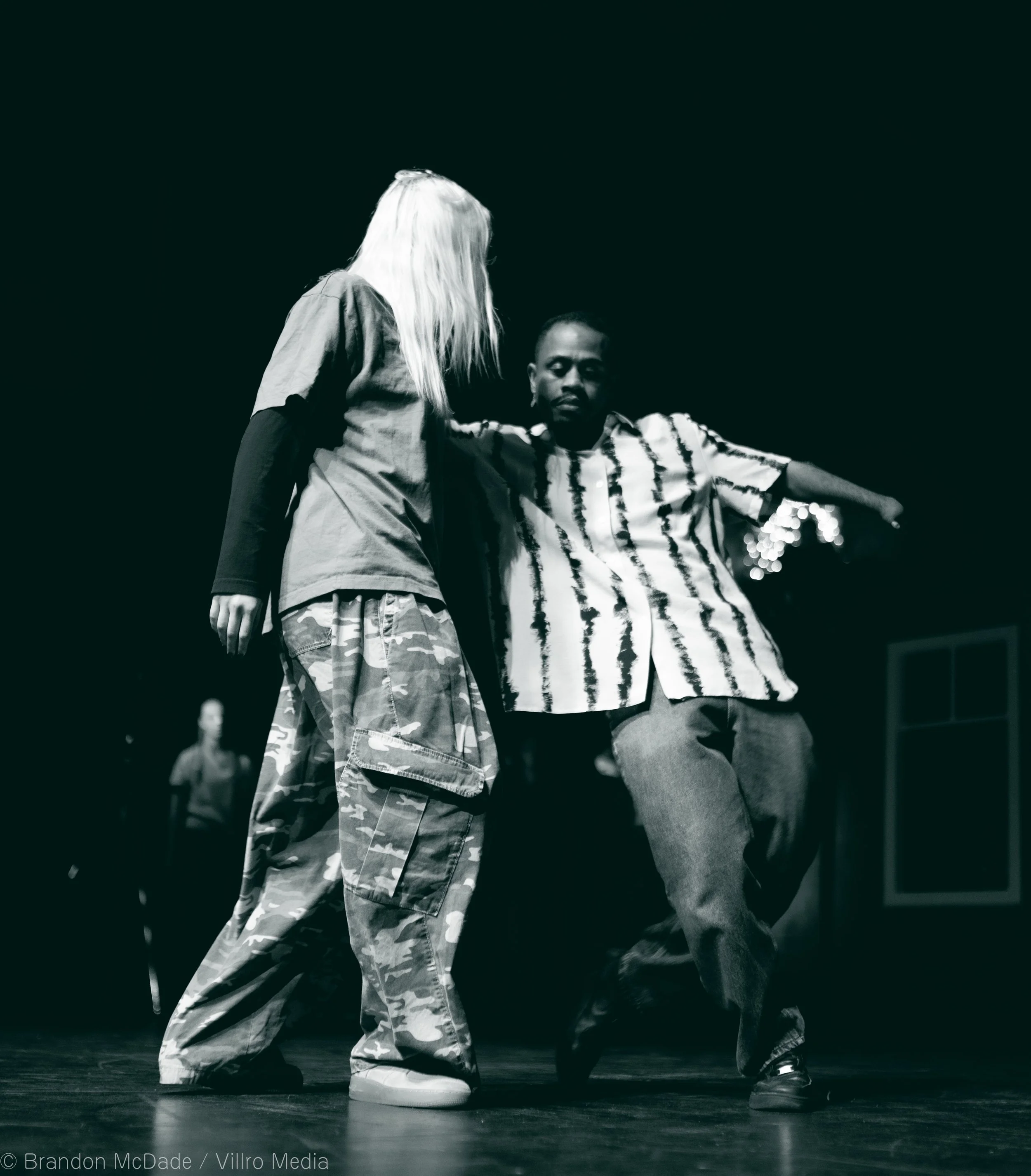 Two people, one in camouflage pants and a jacket, and the other in striped shirt and baggy pants, dance together on a dark stage.