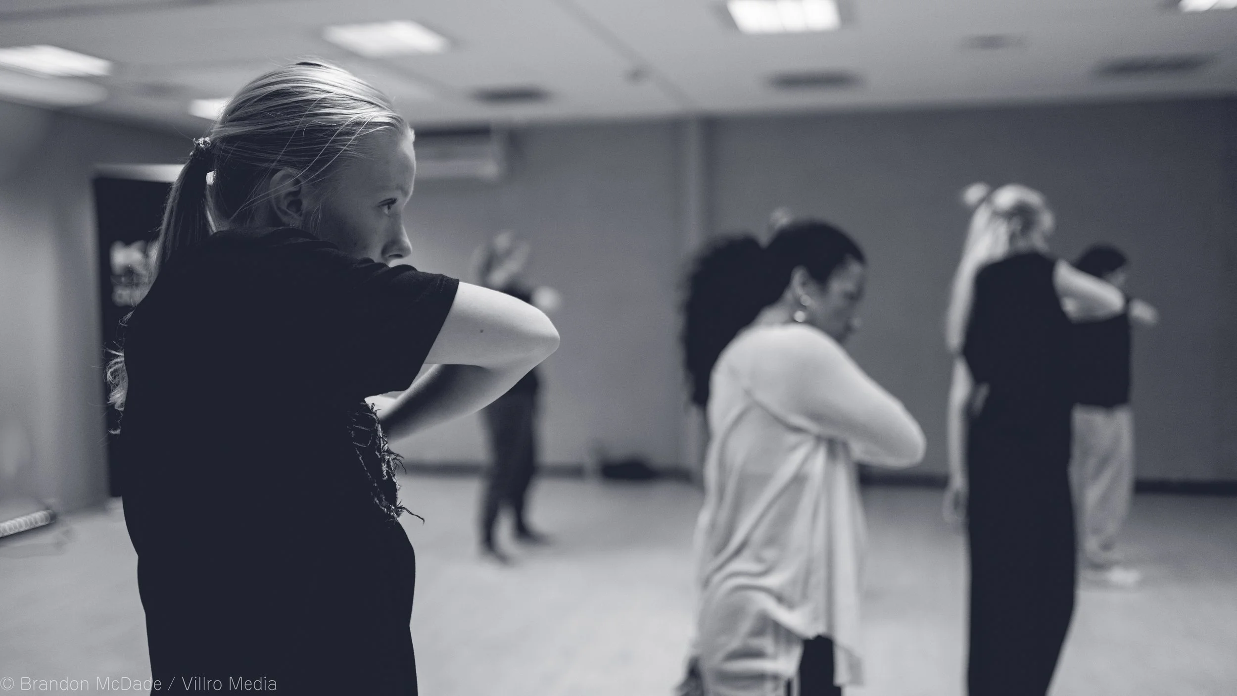 A group of women practicing dance or exercise in a studio, standing in a line with arms crossed or on hips, focused on their movements.
