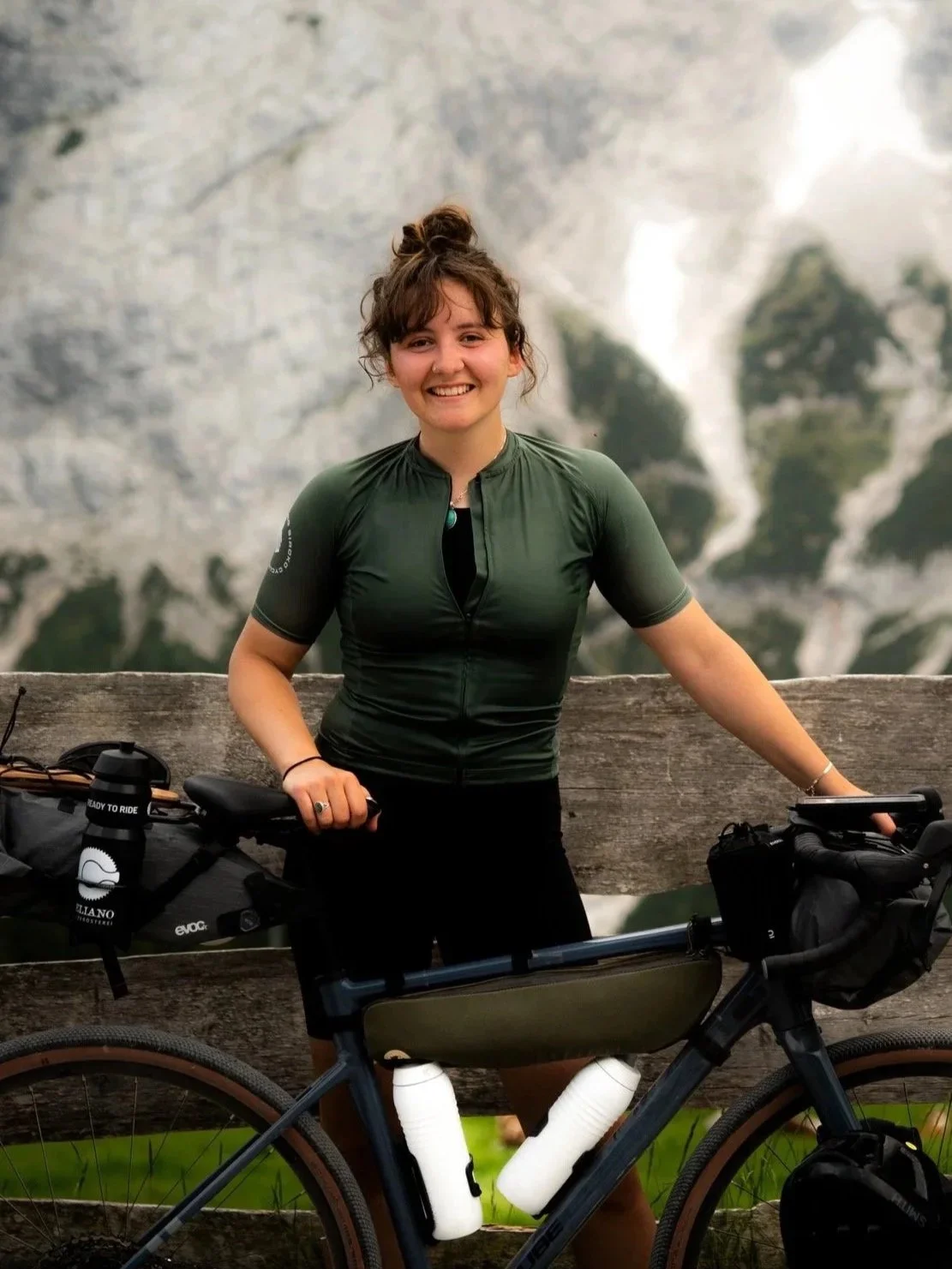 Junge Frau mit einem Fahrrad, die vor einer Holzbarriere steht, trägt grüne Radhandschuhe und hat braune, gewellte Haare, mit einem Berg im Hintergrund.