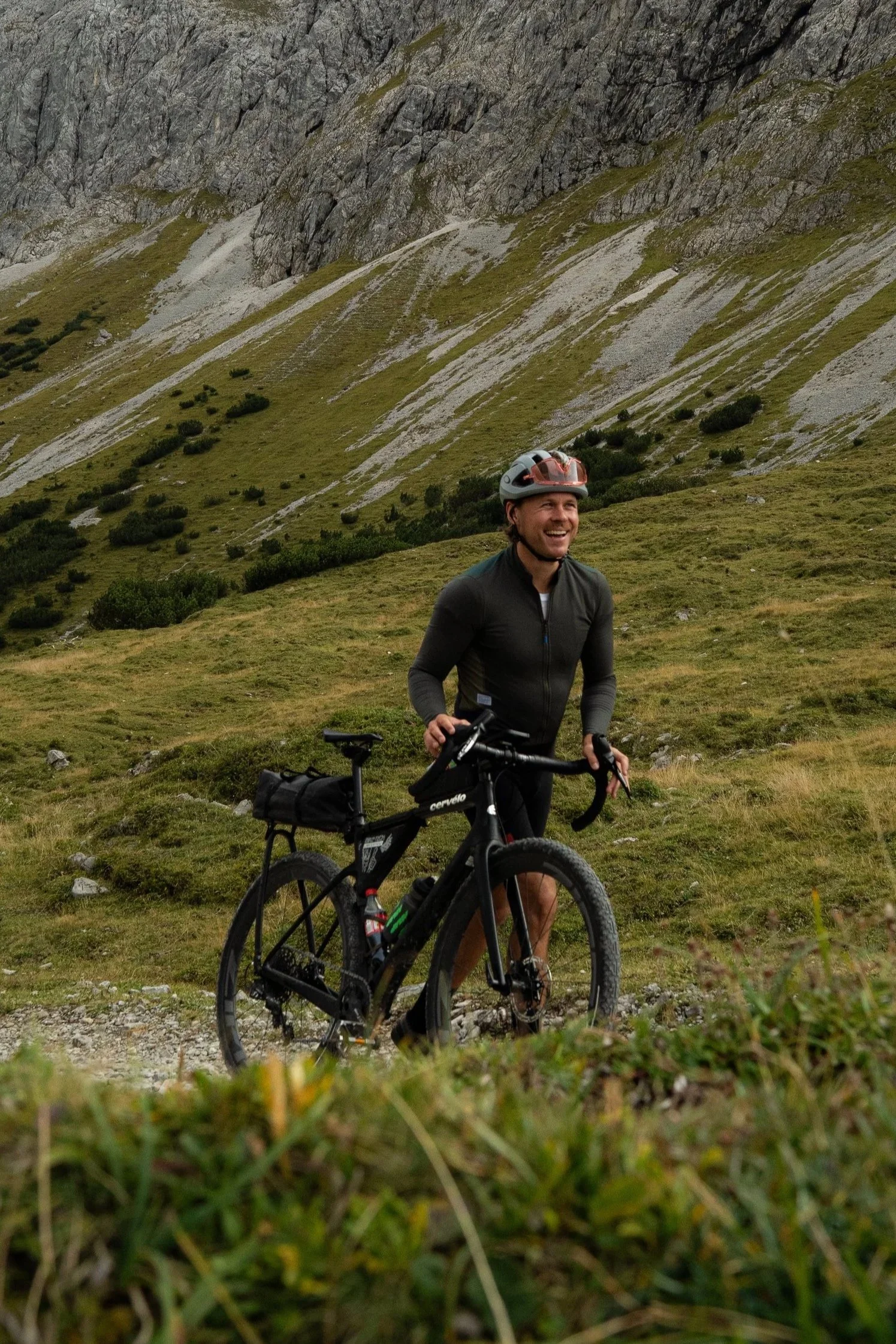 Ein Mann mit Fahrrad in einer bergigen, grünen Landschaft, trägt einen Helm und lächelt.