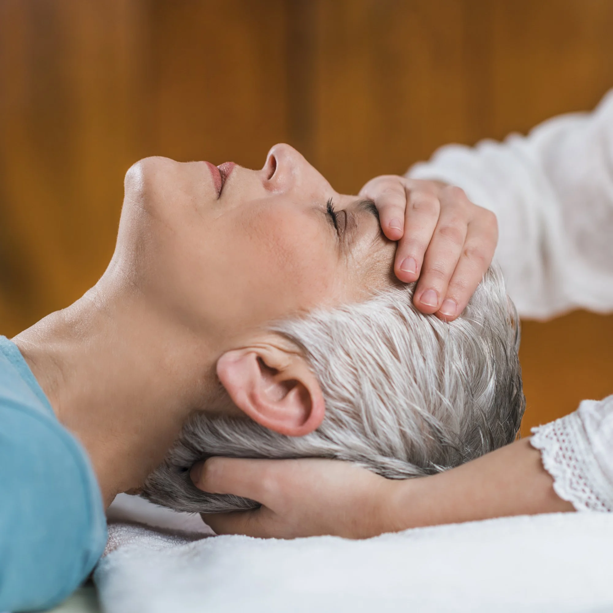 A woman receives a head massage or chiropractic treatment from a person with hands on her forehead and neck, lying on a white surface with a wooden background.