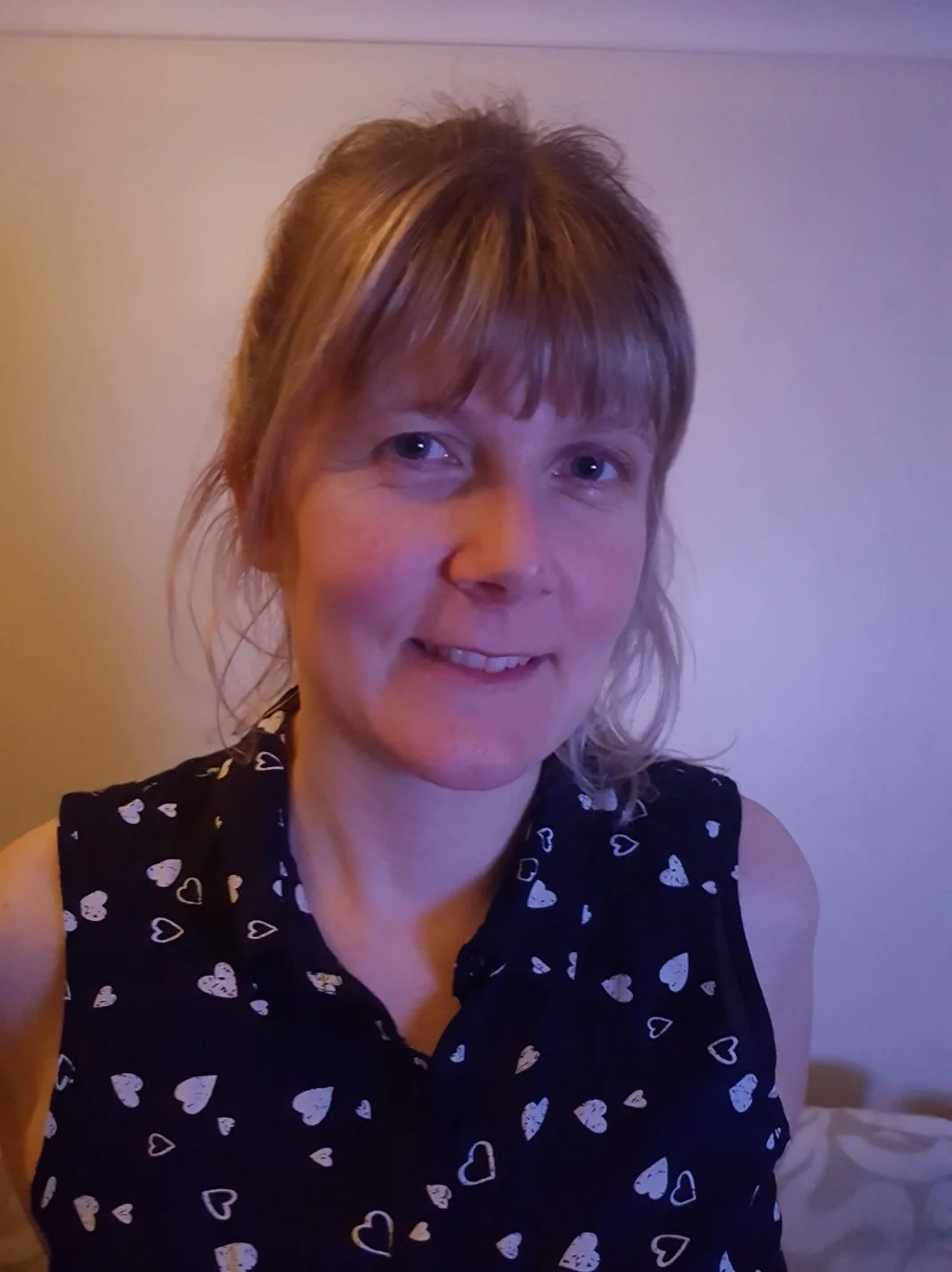 A woman with light brown hair with highlights, cut in a choppy style with bangs, wearing a black sleeveless top with white heart patterns, smiling slightly, in a room with plain light-colored walls.