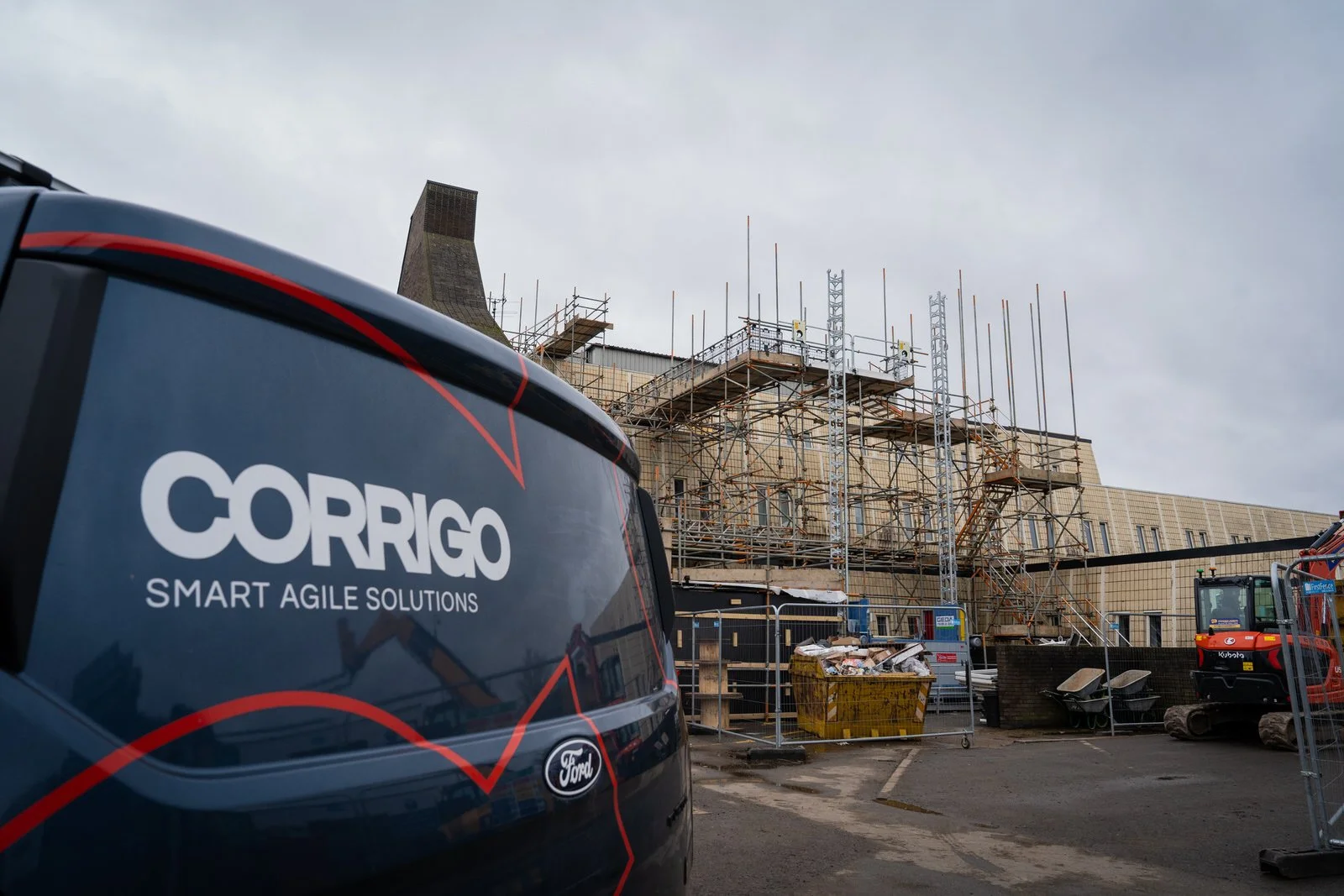 Construction site with scaffolding around a building, construction equipment, a yellow dumpster filled with debris, and a black vehicle with the logo 'CORRIG SMART AGILE SOLUTIONS' in the foreground.