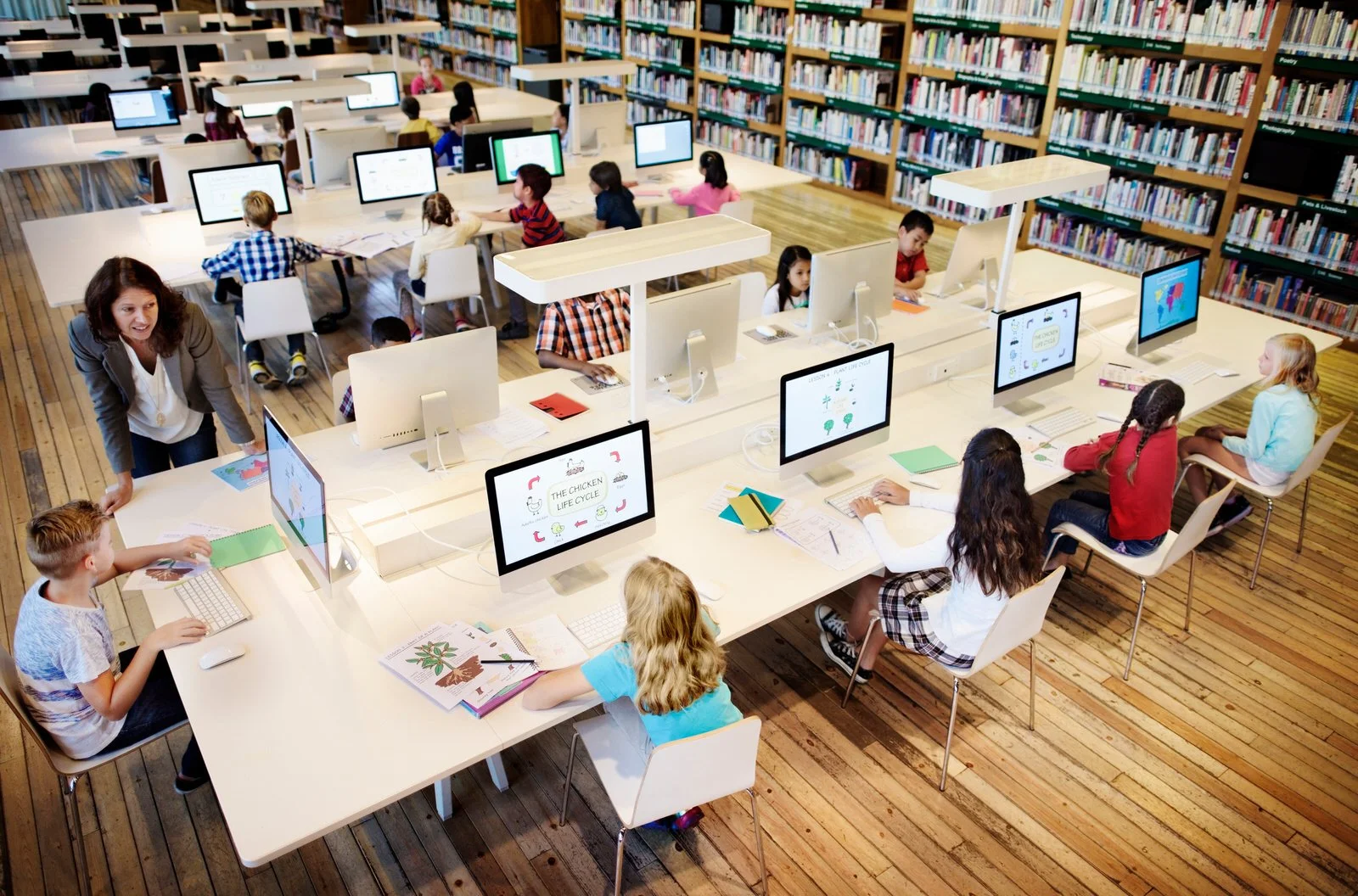 Children working on computers in a school library with bookshelves in the background.