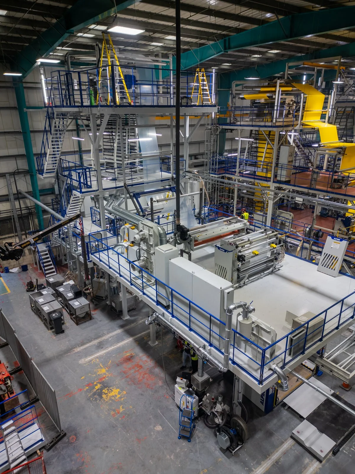 Inside a factory with large industrial machinery, pipes, stairs, and walkways, with a few workers wearing safety gear.