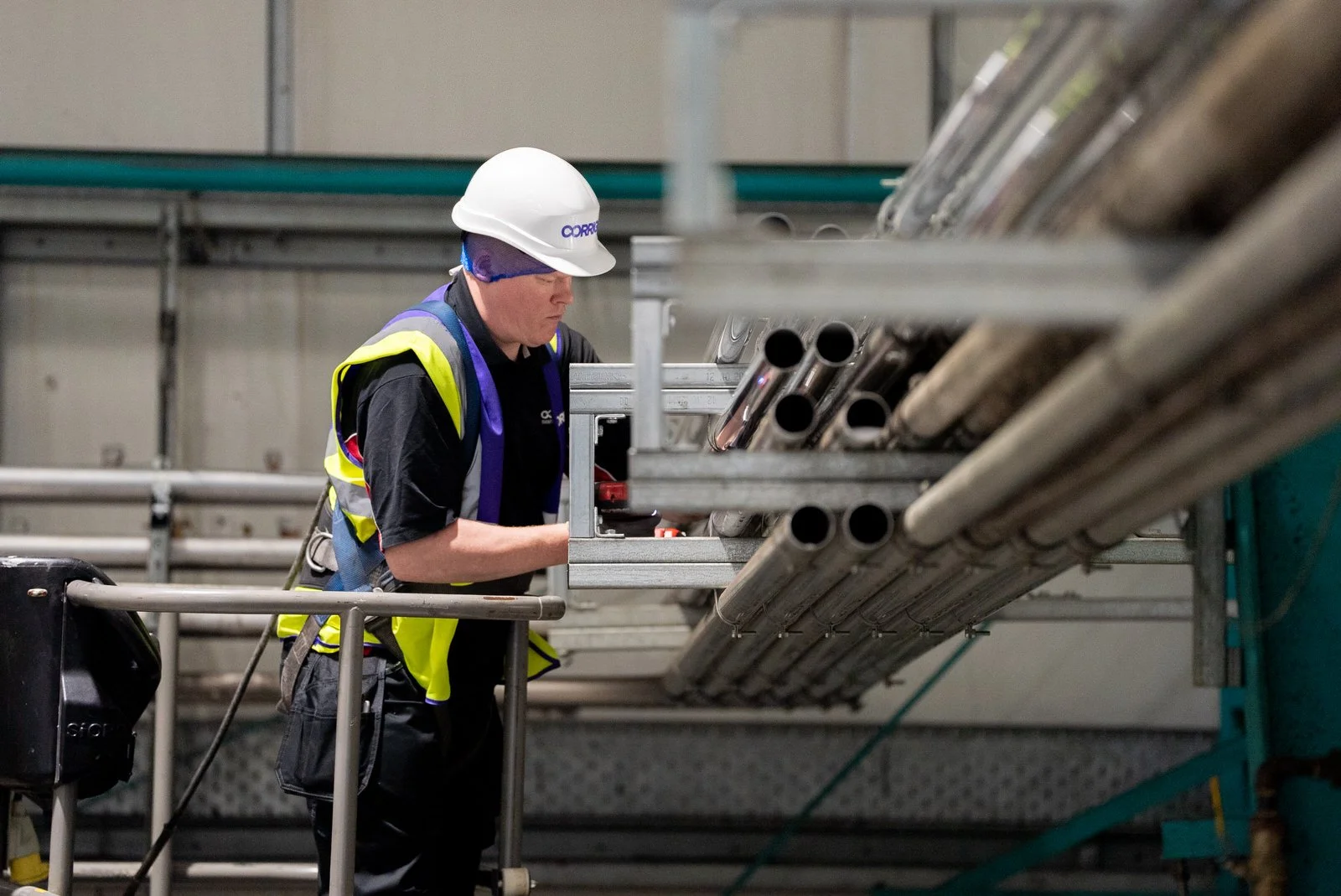A worker wearing a white hard hat, yellow safety vest, and black clothing is examining metal pipes on a metal rack inside an industrial facility.