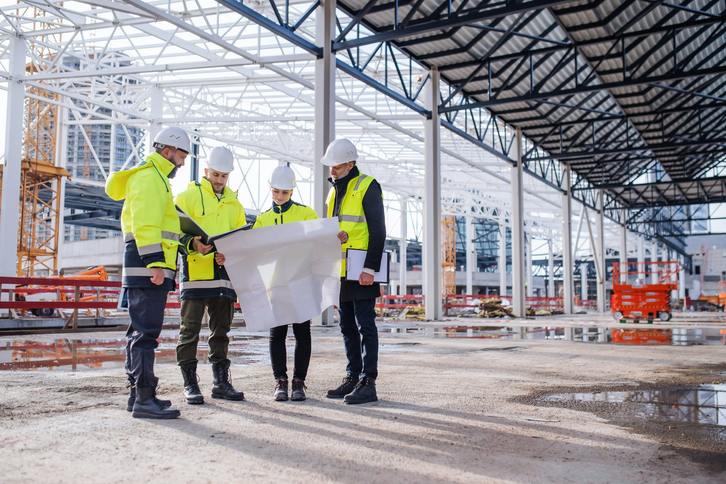 Construction workers wearing safety vests and helmets examining blueprints on a construction site with steel framework and a building under construction.