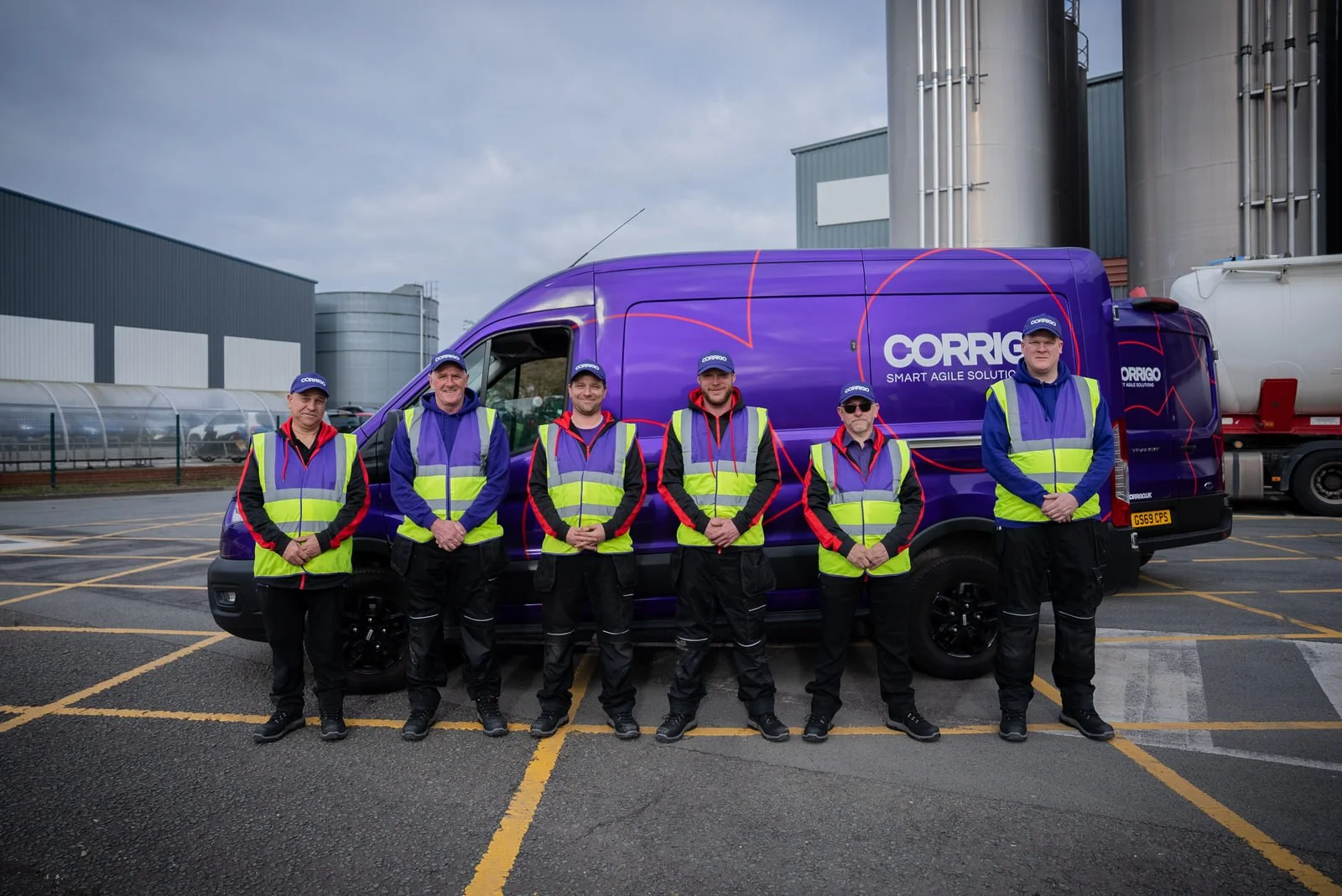 Six men standing in front of a purple van with a logo that reads "CORRIGO SMART AGILE SOLUTIONS," all wearing safety vests and caps, in an outdoor parking lot near industrial buildings.