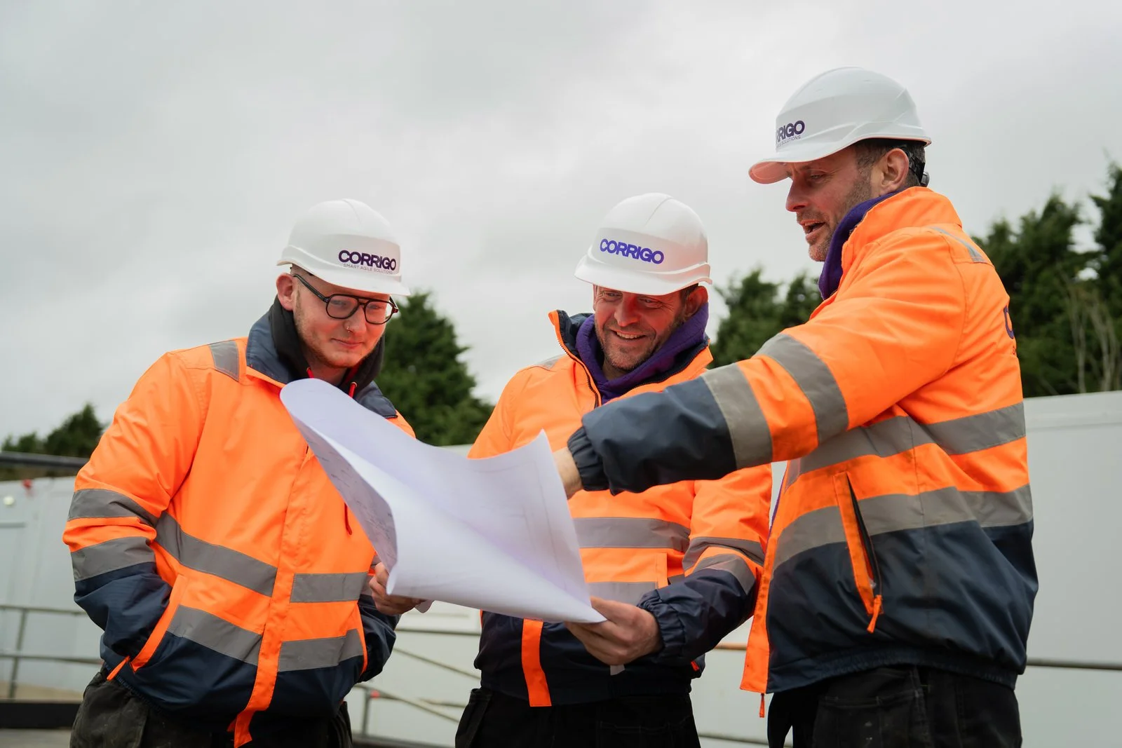 Three construction workers wearing white helmets and orange high-visibility jackets standing outdoors, reviewing blueprints and discussing plans.