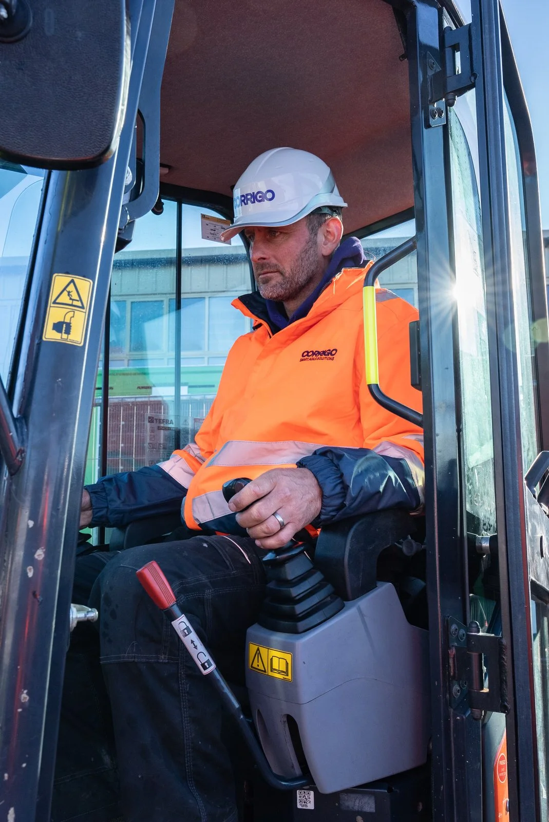 A man in an orange safety jacket and white hard hat operating a piece of construction equipment, possibly a skid steer loader, inside a cabin.