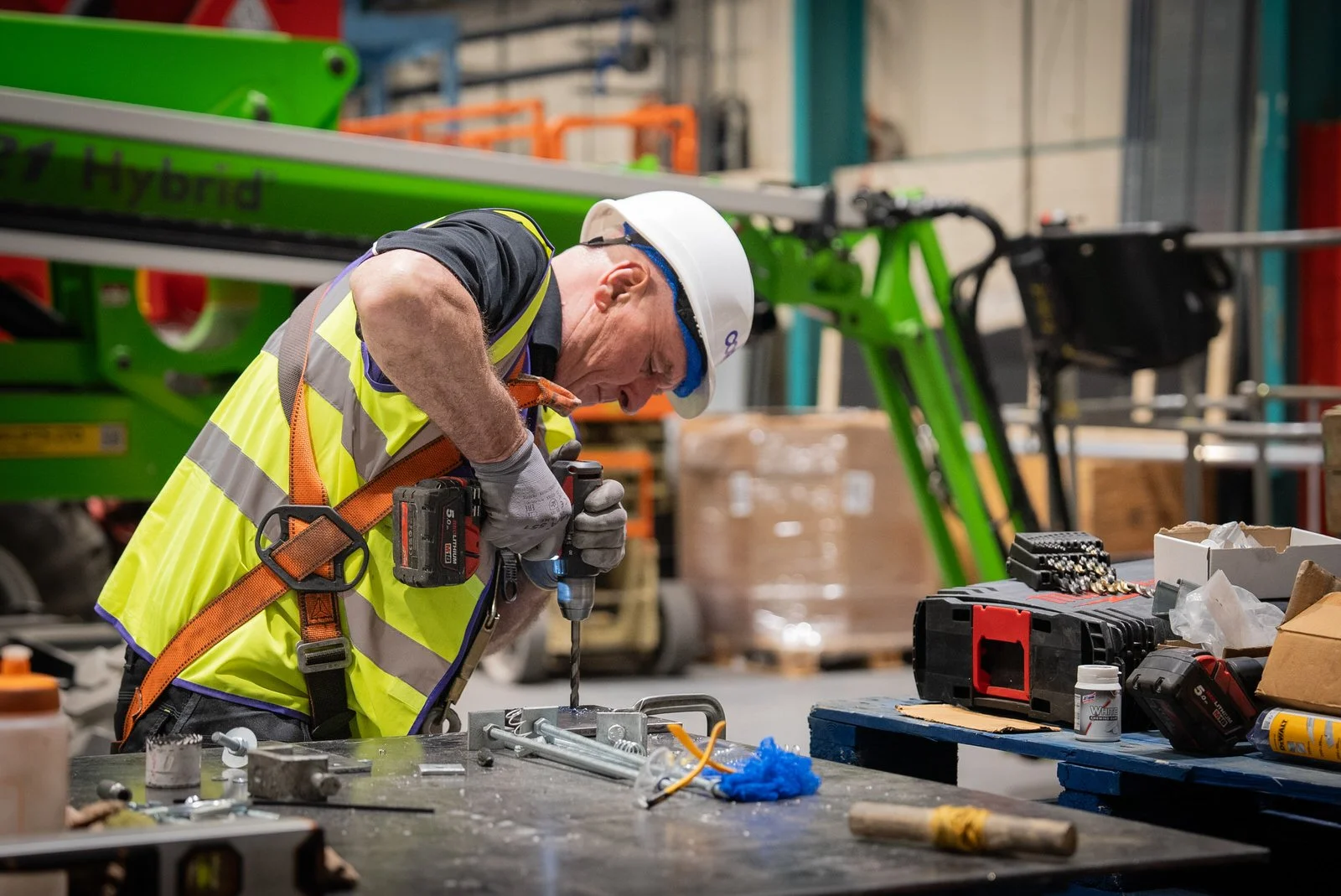 A worker in a yellow safety vest and white hard hat using a power drill at a workbench in an industrial warehouse surrounded by tools and equipment.
