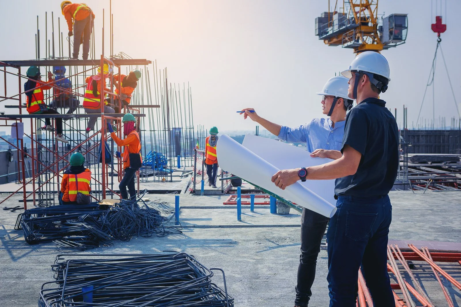 Construction workers wearing safety vests and helmets working on site with scaffolding, rebar, and construction equipment, and two supervisors in helmets reviewing plans.