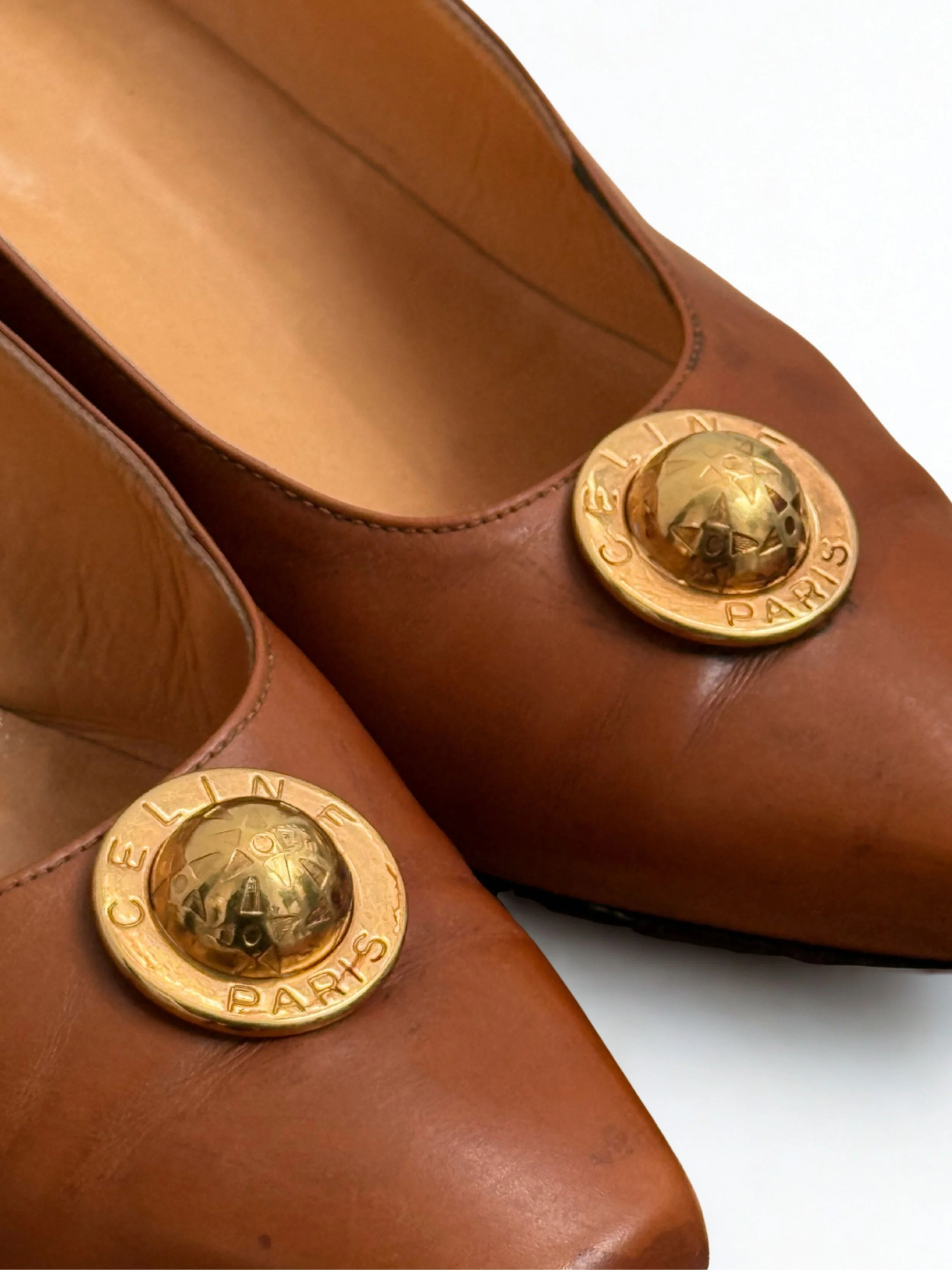 A pair of brown Céline leather shoes with gold-toned embellishments on top. The embellishments read 'Céline PARIS' and are spherical, resembling a globe.