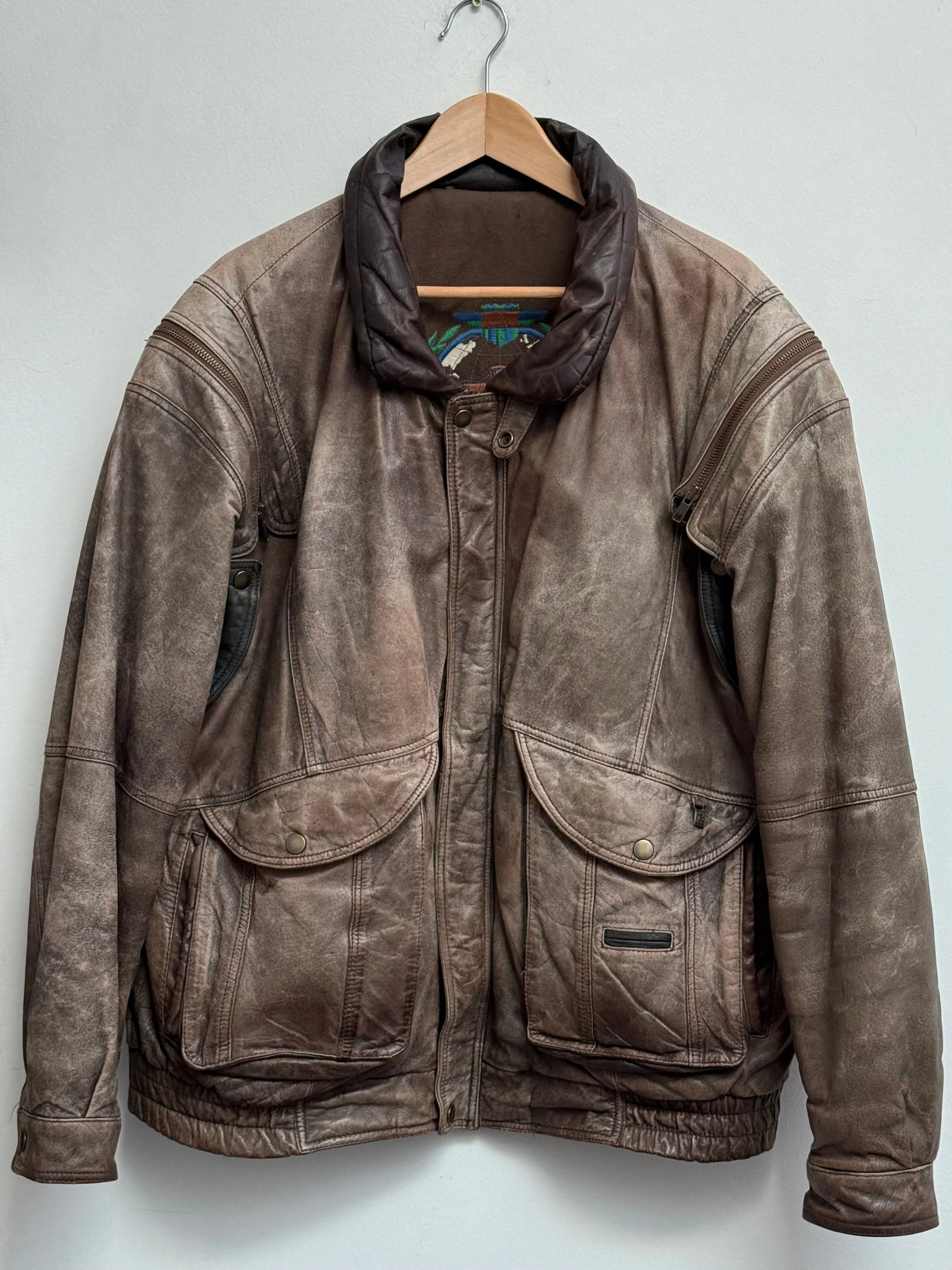 A vintage brown leather bomber jacket hanging on a wooden hanger against a plain white wall.