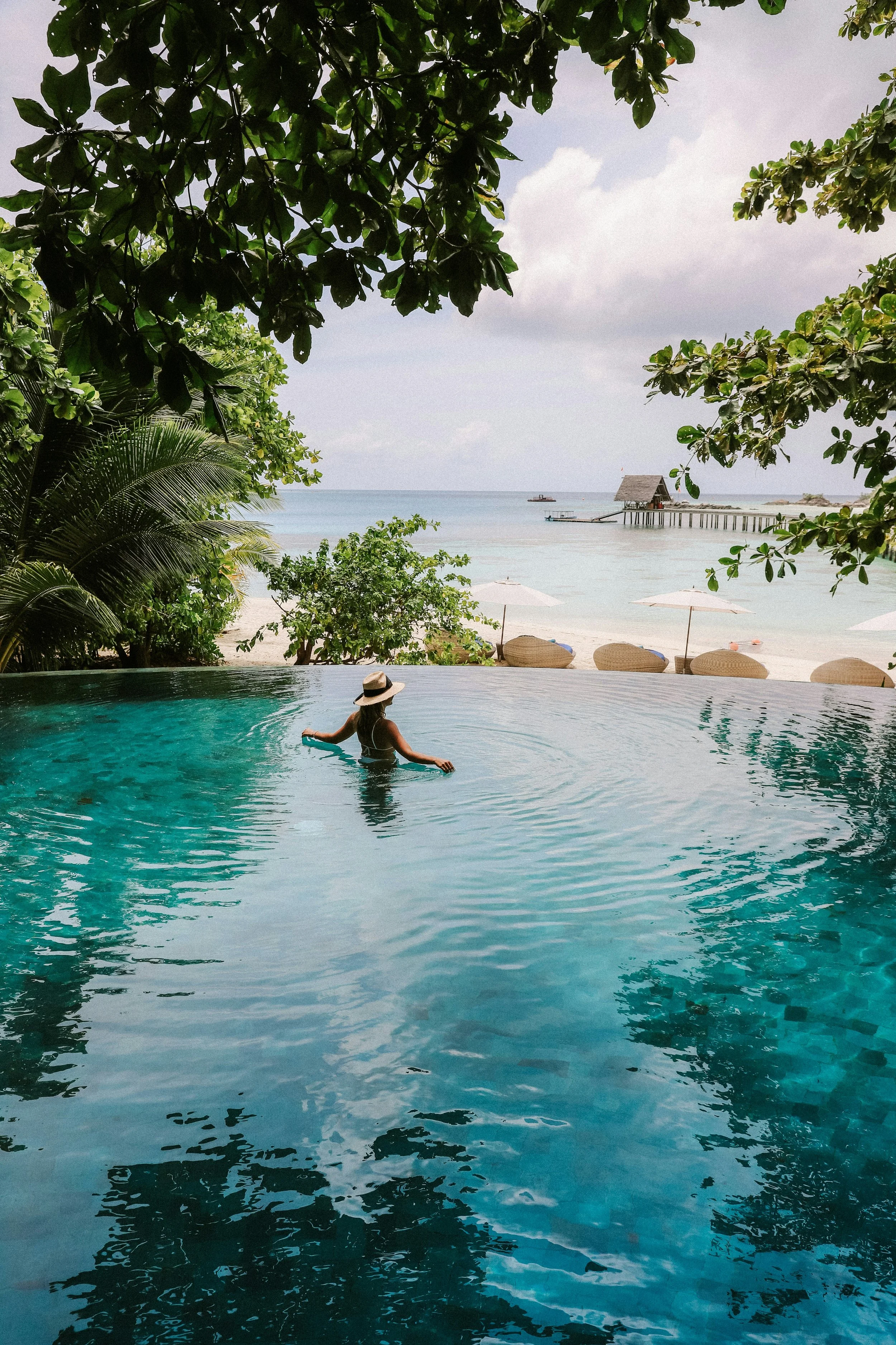A person in a hat enjoying an infinity pool overlooking a tropical beach and ocean with a pier in the distance, framed by leafy green trees.