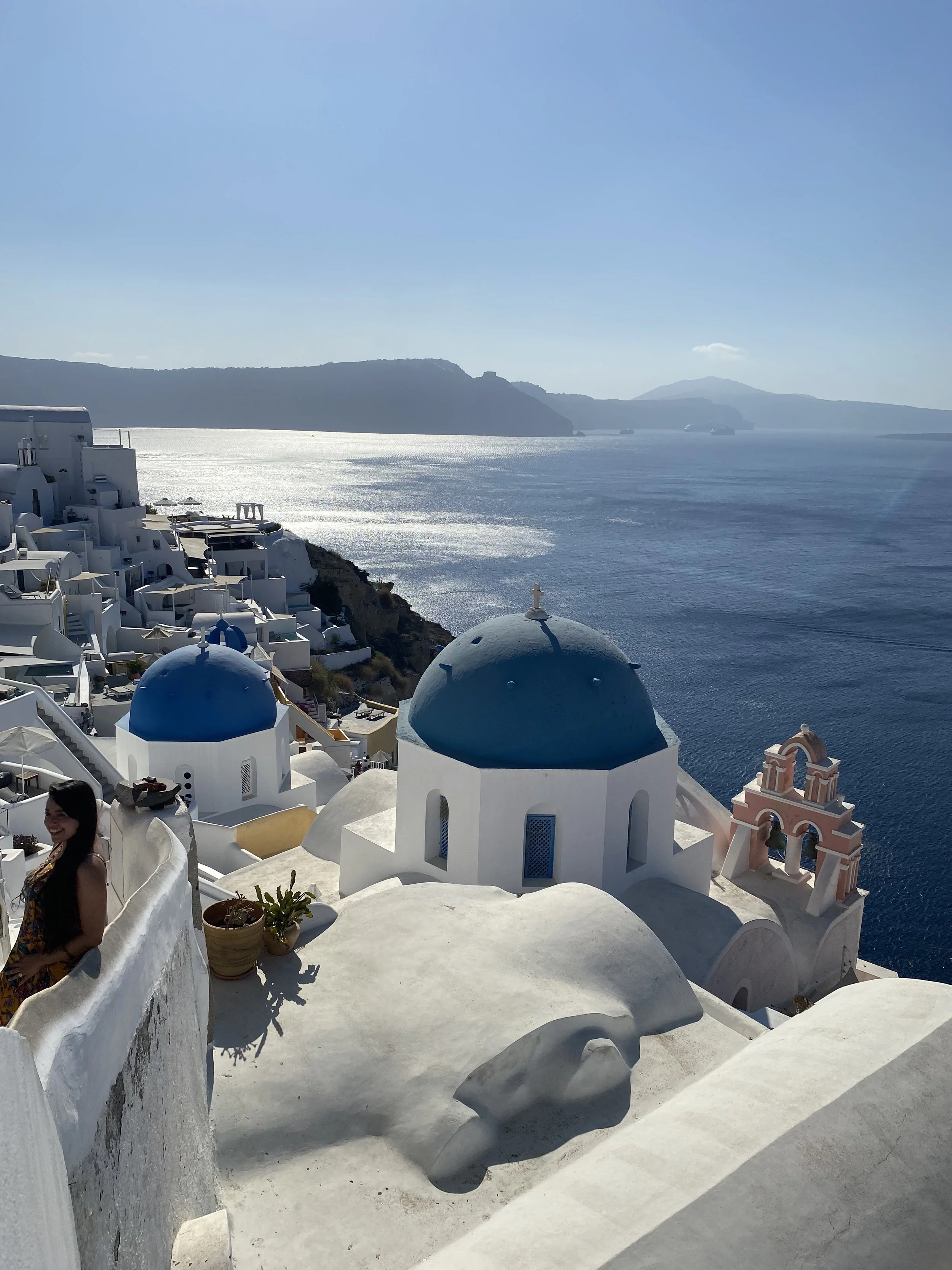 Scenic view of white buildings with blue domes overlooking the sea in Santorini, Greece, with a distant mountainous island and clear blue sky.