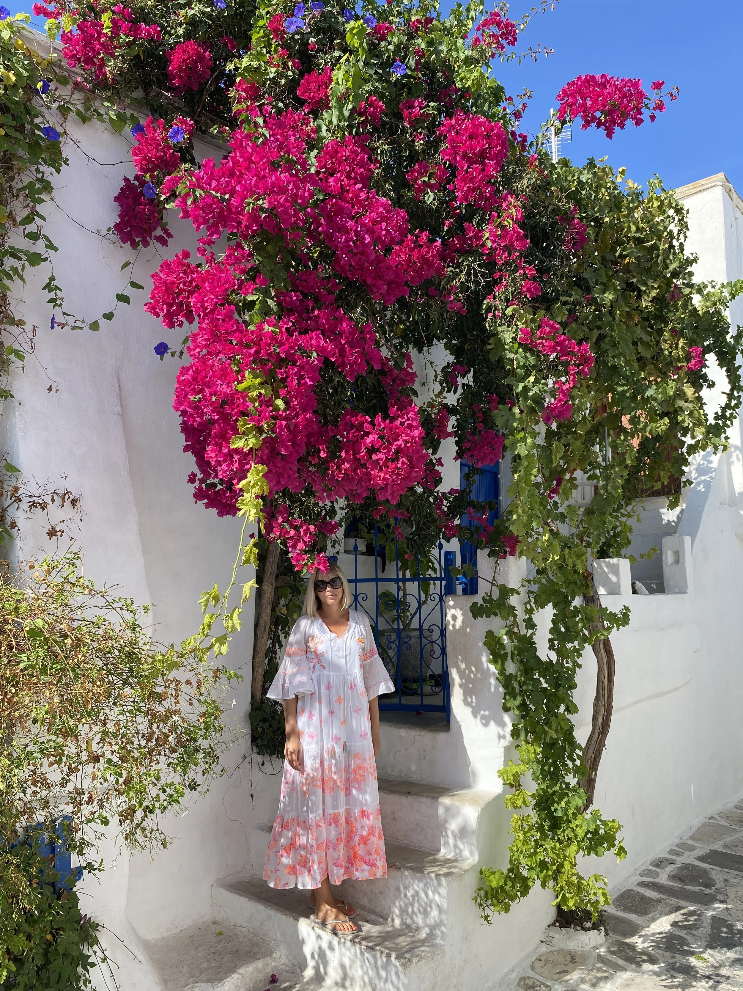 A woman in a long white dress with pink and orange floral patterns standing on white stone steps, surrounded by bright pink bougainvillaea flowers and green foliage, with a blue door and iron gate in the background in Paros Greece