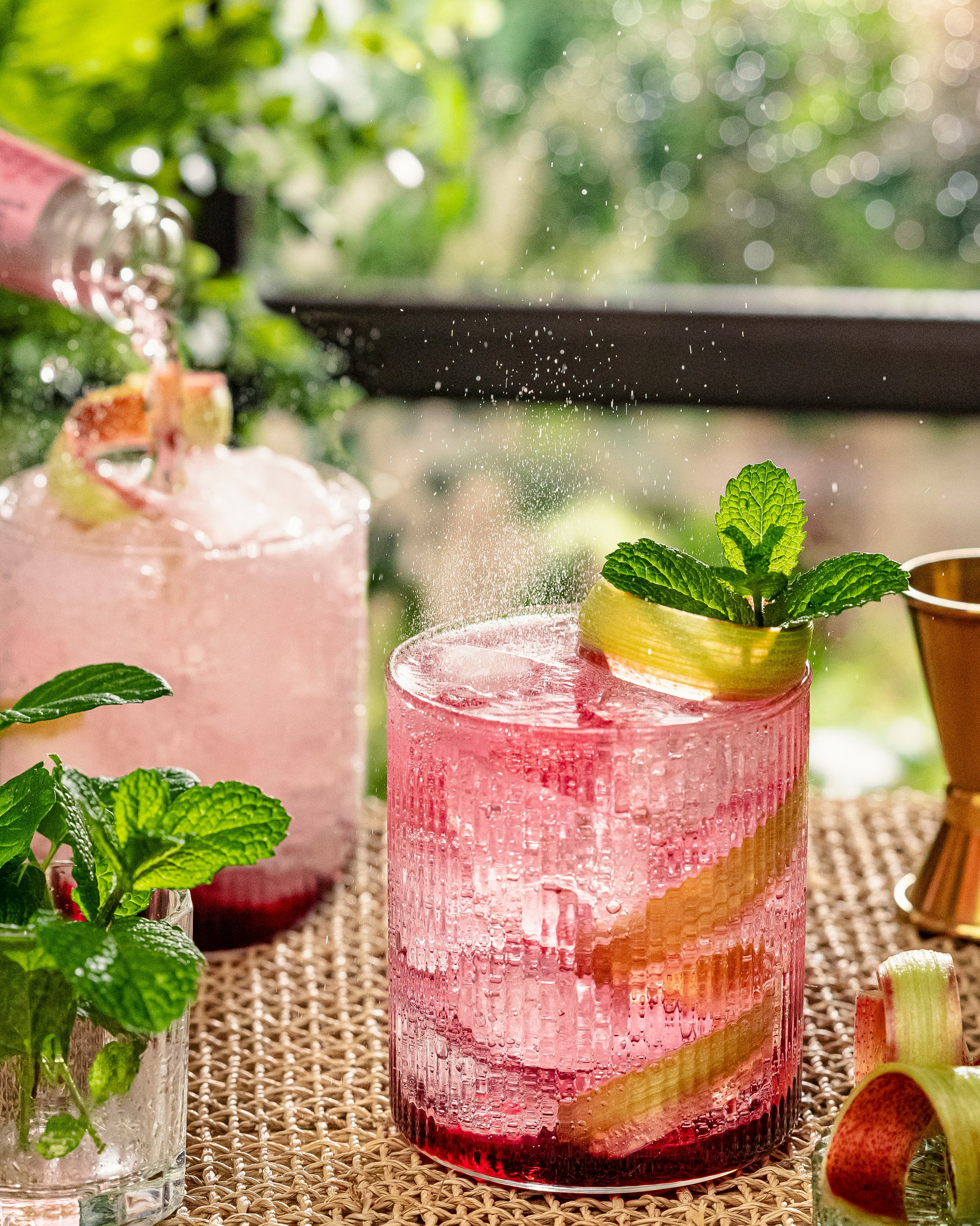 A pink cocktail garnished with a slice of cucumber and fresh mint leaves. In the background, there is another similar drink and a small pot of mint leaves on a woven mat, with a blurred green outdoor scene.
