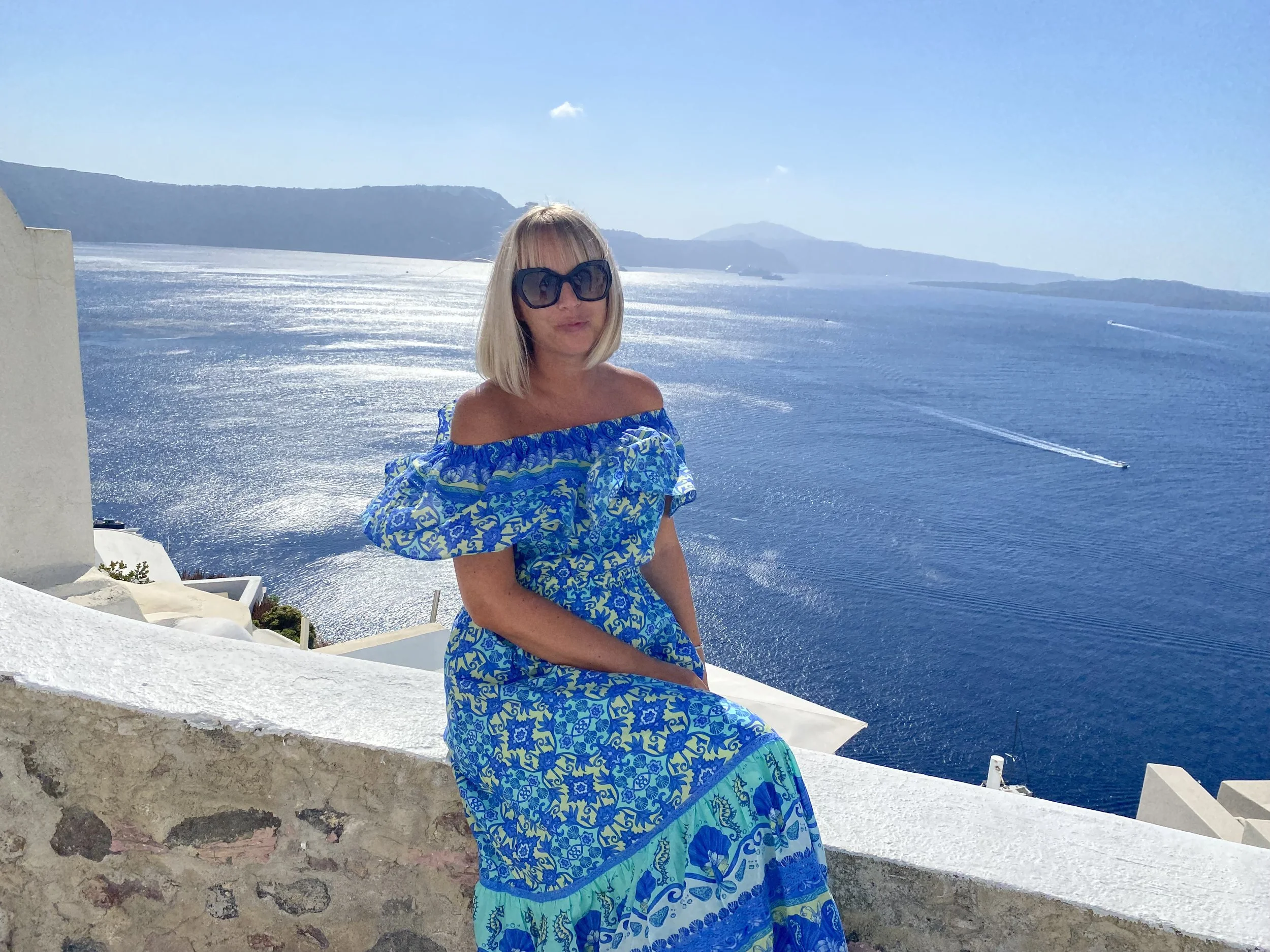 A woman with blonde hair wearing oversized sunglasses and a blue off-shoulder dress sitting on a white stone ledge overlooking a large body of water with boats in Santorini