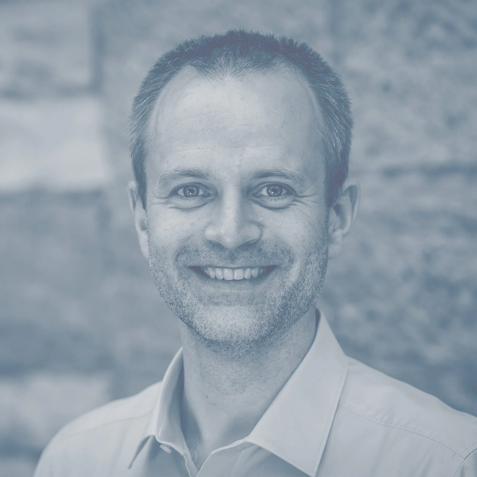 Nahaufnahme eines lächelnden Mannes mit kurzbraunem Haar vor einem Steinmauerhintergrund.