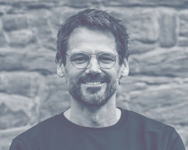 Ein lächelnder Mann mit Brille, dunklem T-Shirt und Bart vor einer Steinmauer.