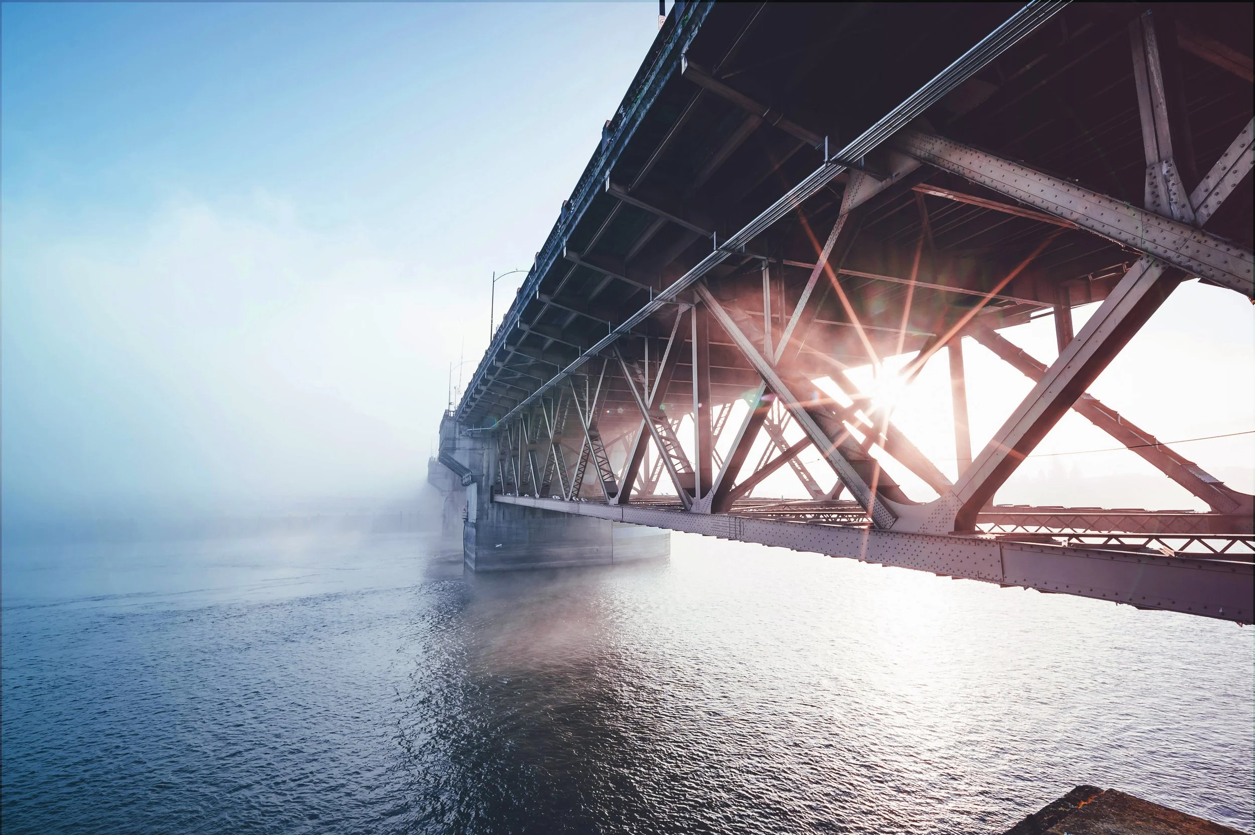 Blick unter einer Brücke über Wasser, Sonnenstrahlen scheinen durch die Brückenstruktur, im Morgennebel.