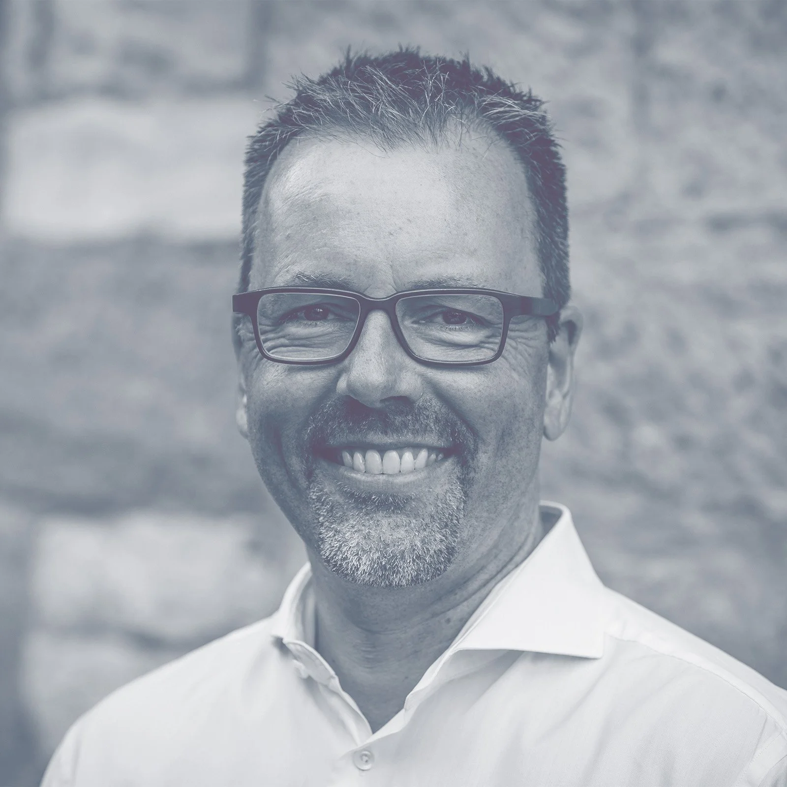 Ein lächelnder Mann mit Brille und kurzem, spitzigem Haar vor einer Backsteinmauer.