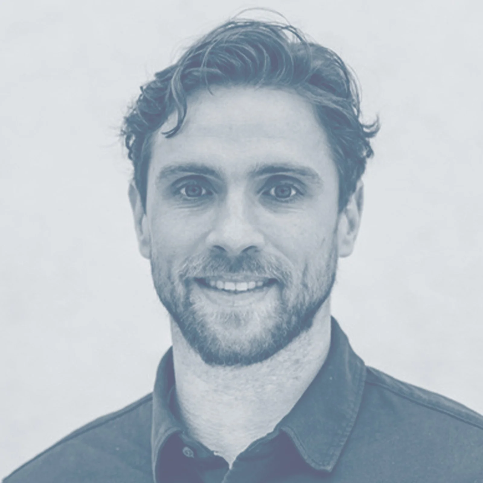 Schwarz-weiß Porträt eines lächelnden Mannes mit kurzen, gewellten Haaren und Bart, trägt ein dunkles Hemd, vor hellem Hintergrund.