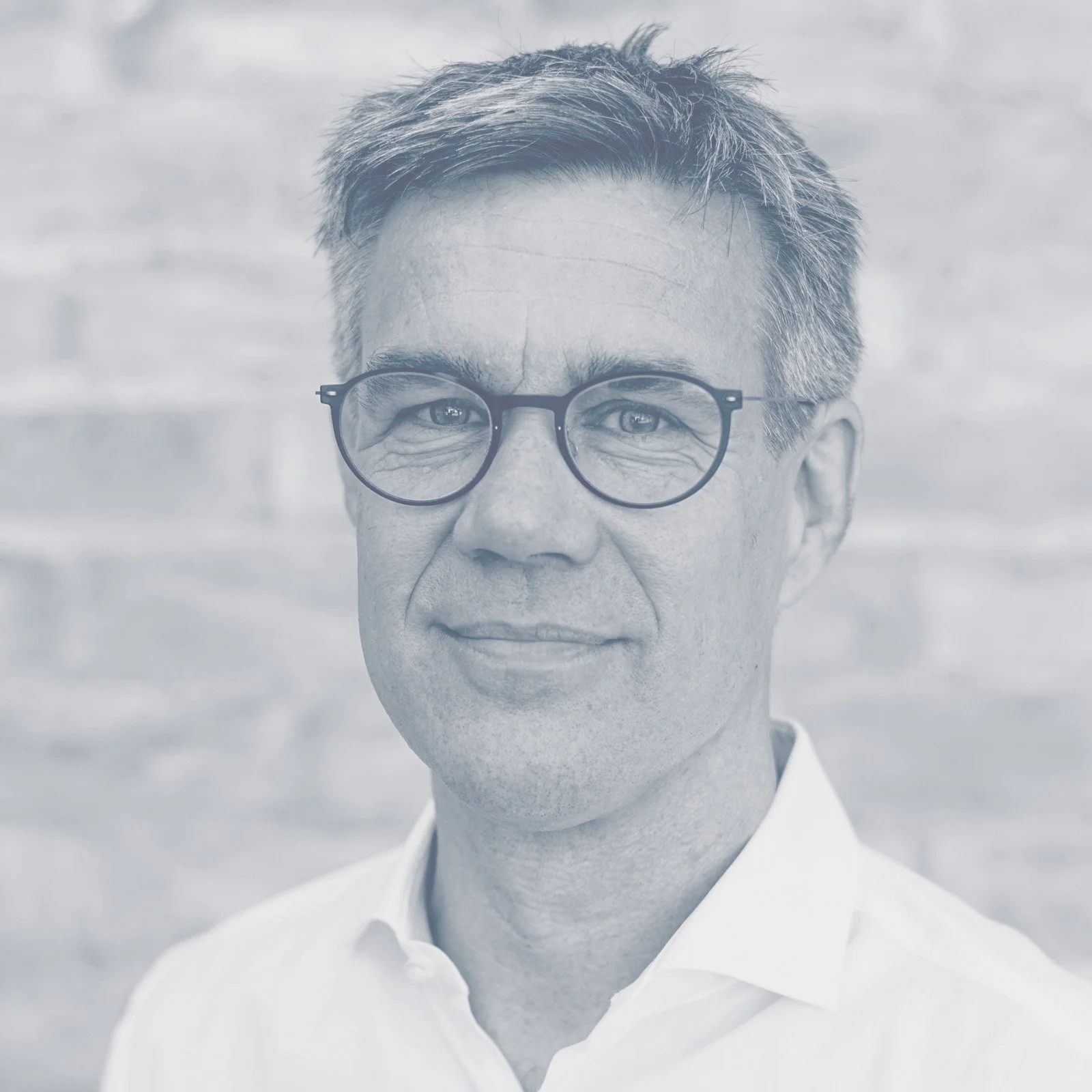 Porträt eines Mannes mit Brille und weißem Hemd vor einer hellen, strukturierten Wand.